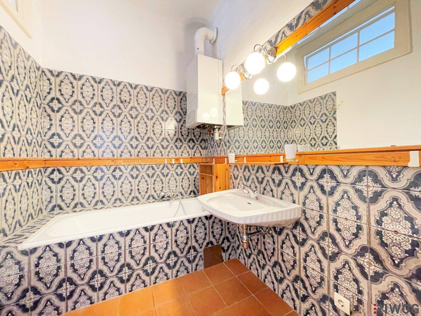 Badezimmer mit gemusterten Fliesen, Badewanne und Waschbecken unter einem Fenster.