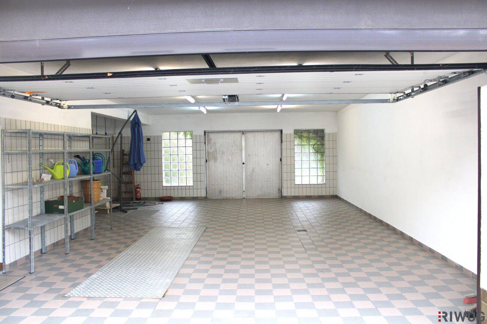 Geräumige Garage mit gefliestem Boden, Regalen und zwei großen Toren im hinteren Bereich des Raumes.