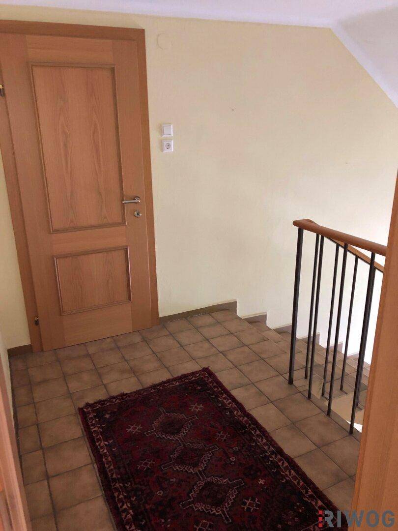 Eingangsbereich mit Treppe, gefliestem Boden und einer Holztür, die ins Innere des Hauses führt.
