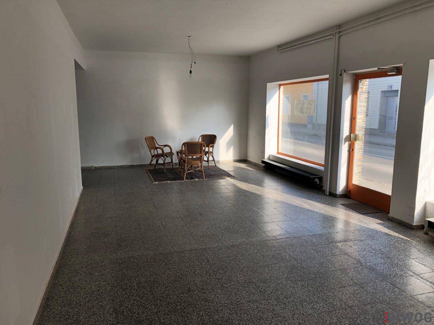 Leerer Geschäftsraum mit großen Schaufenstern zur Straße, gefliestem Boden und einigen Korbstühlen.