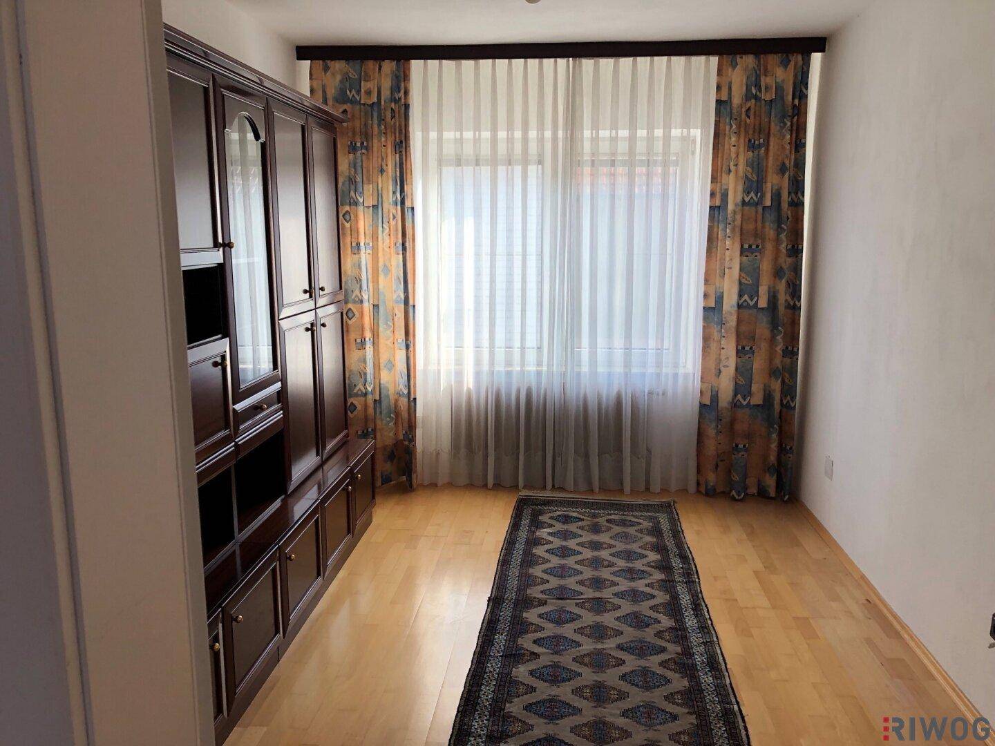 Wohnraum mit dunklem Holzschrank, Parkettboden und großen Fenstern mit Vorhängen.
