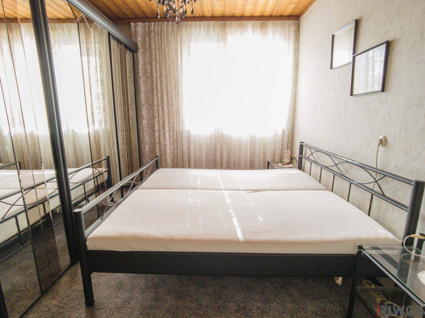 Schlafzimmer mit Doppelbett, Spiegelschrank und Holzdecke, hell durch großes Fenster.