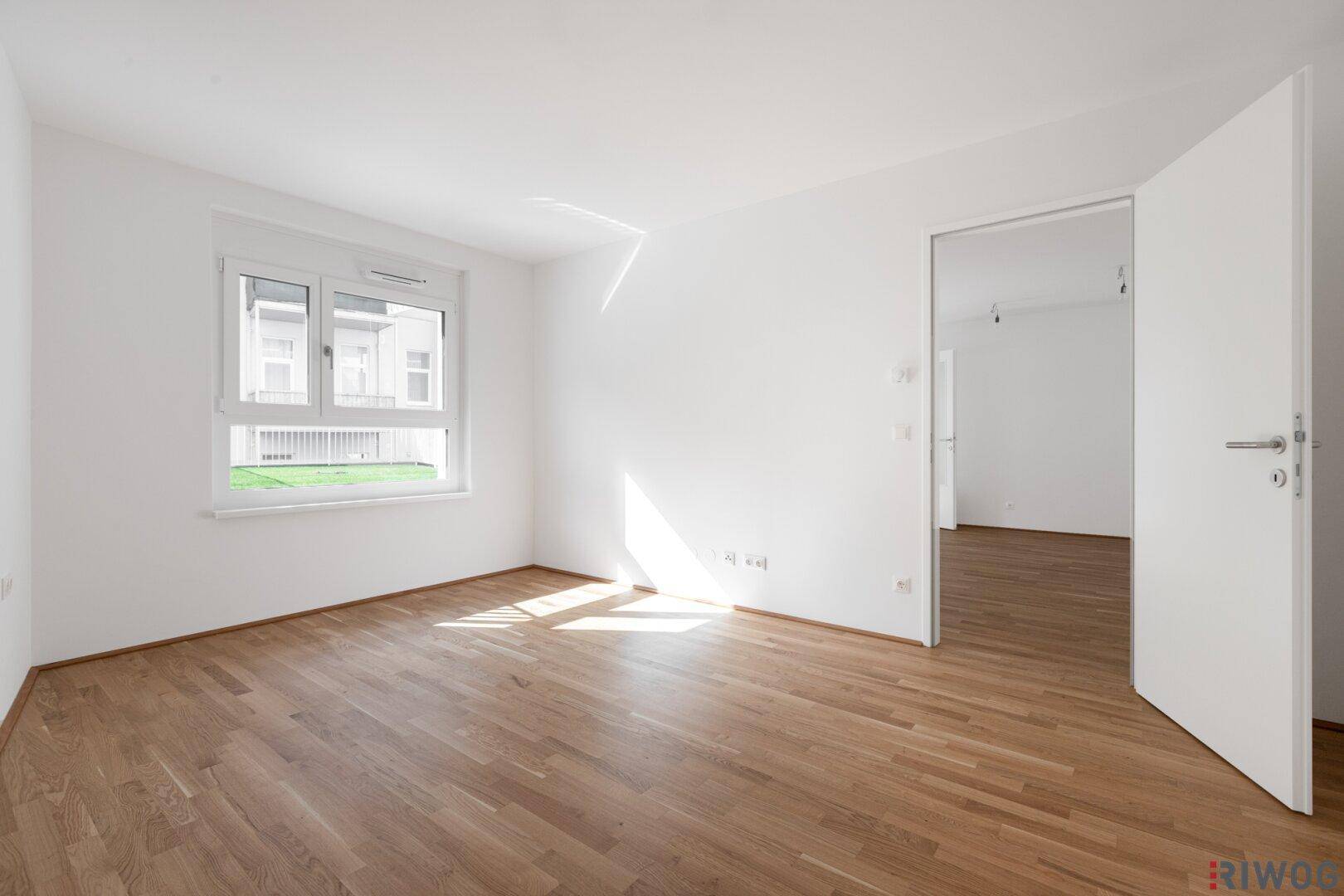 Sonniges Schlafzimmer mit hellem Parkettboden und großen Fenstern, die viel Tageslicht hereinlassen.