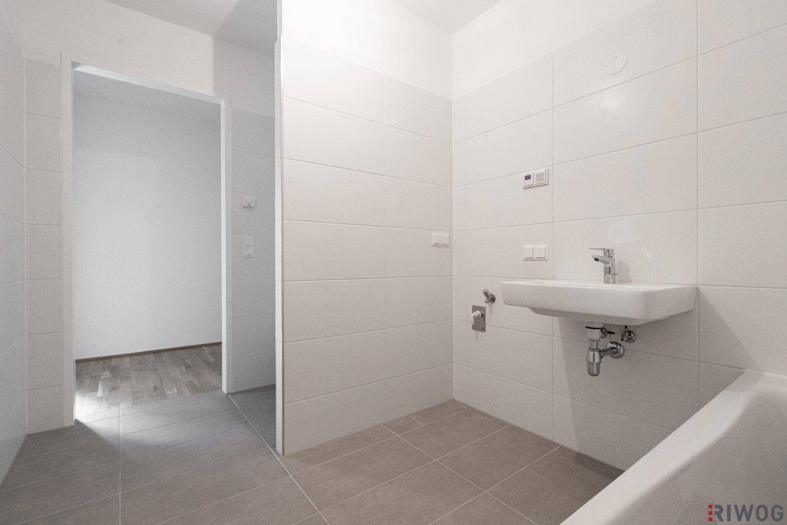 Helles Badezimmer mit weißen Wandfliesen, Einzelwaschbecken und Badewanne, angrenzend an einen weiteren Raum.