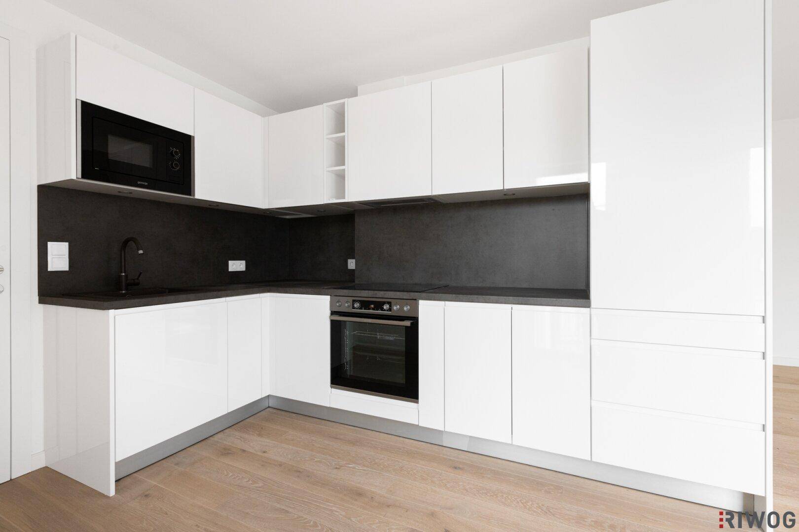 Moderne, weiße Küchenzeile mit glänzenden Fronten, integriertem Backofen und Mikrowelle sowie dunkler Arbeitsplatte.