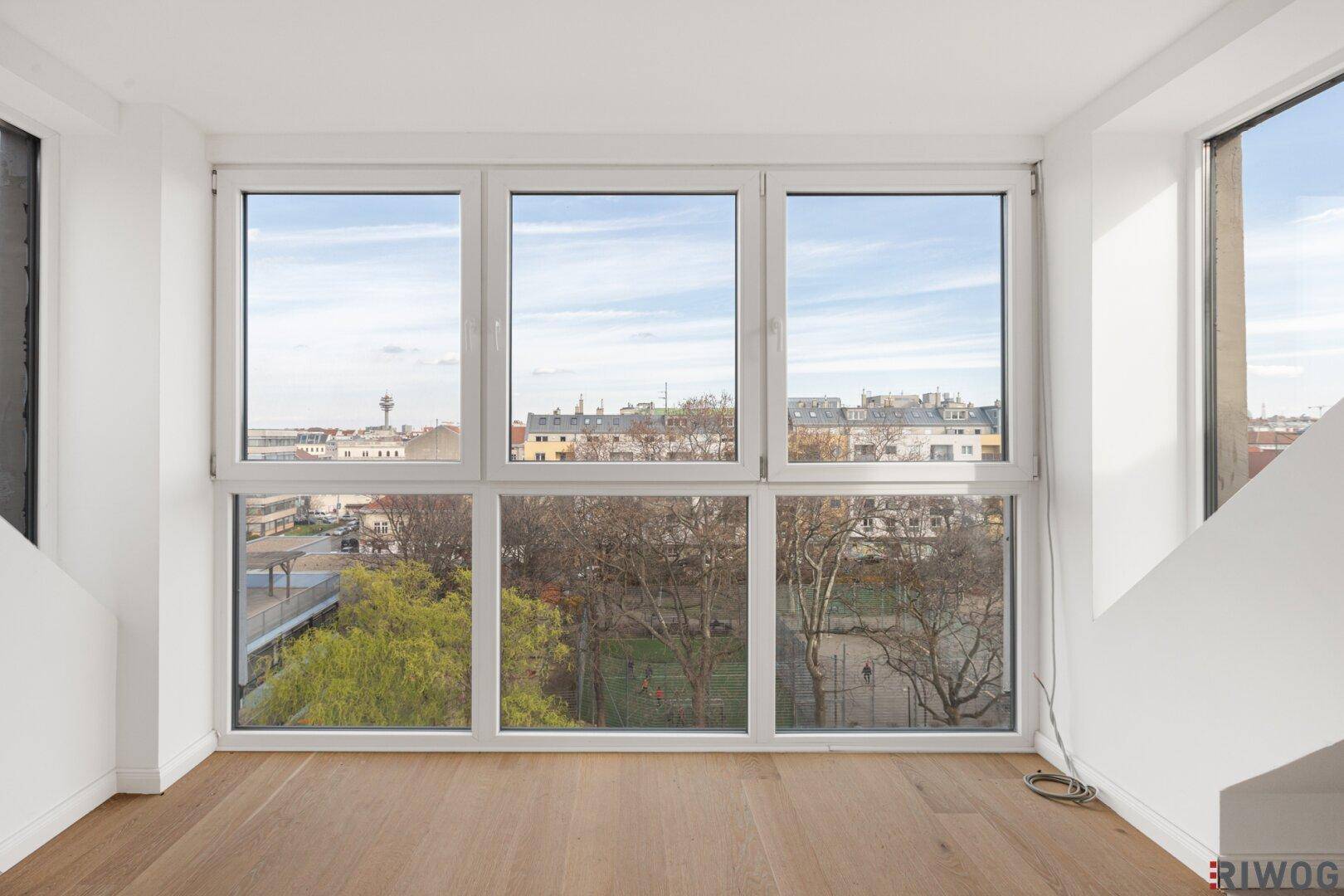 Heller Wohnraum mit großen, bodentiefen Fenstern, Holzboden und weitem Blick über die Stadt und einen Park.