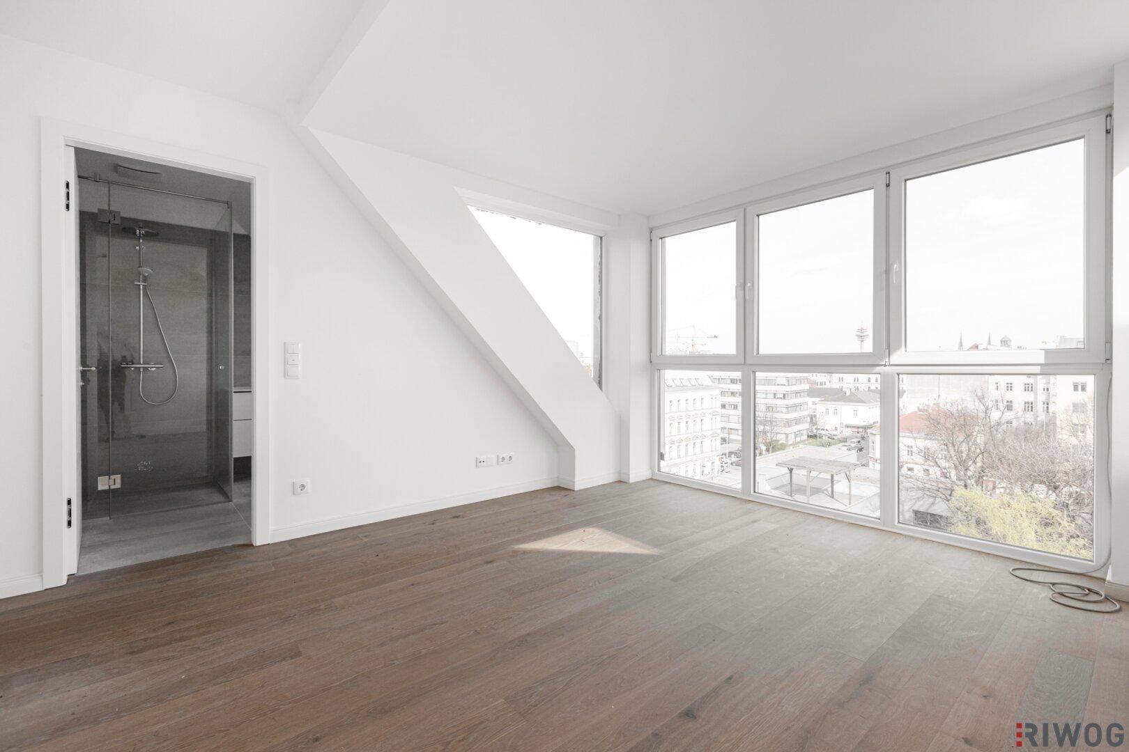 Lichtdurchfluteter Raum mit Holzboden, großen Fenstern und direktem Zugang zu einem modernen Badezimmer.