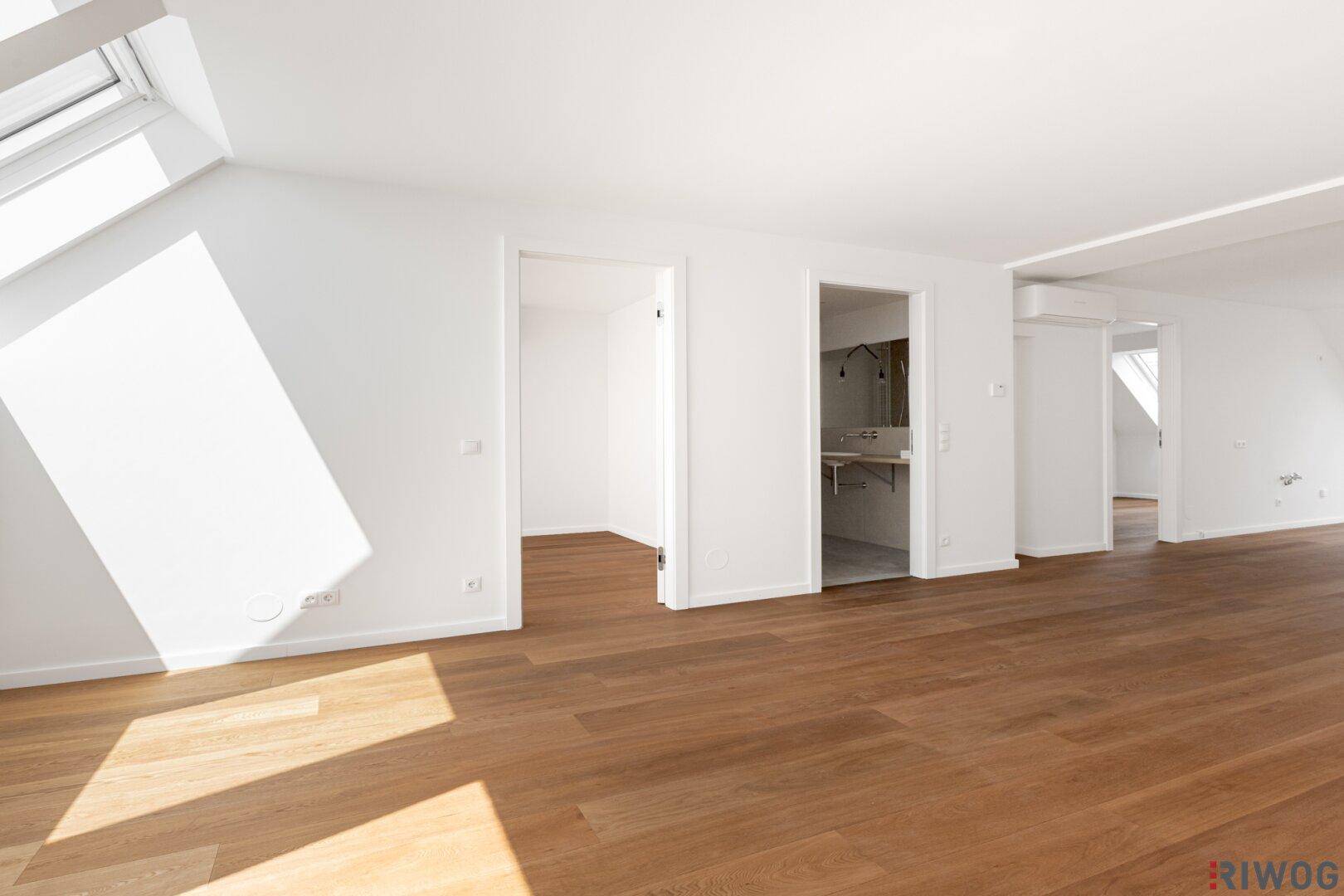 Heller, leerer Raum mit Holzboden und Dachfenstern, der zu einem modernen Badezimmer führt.