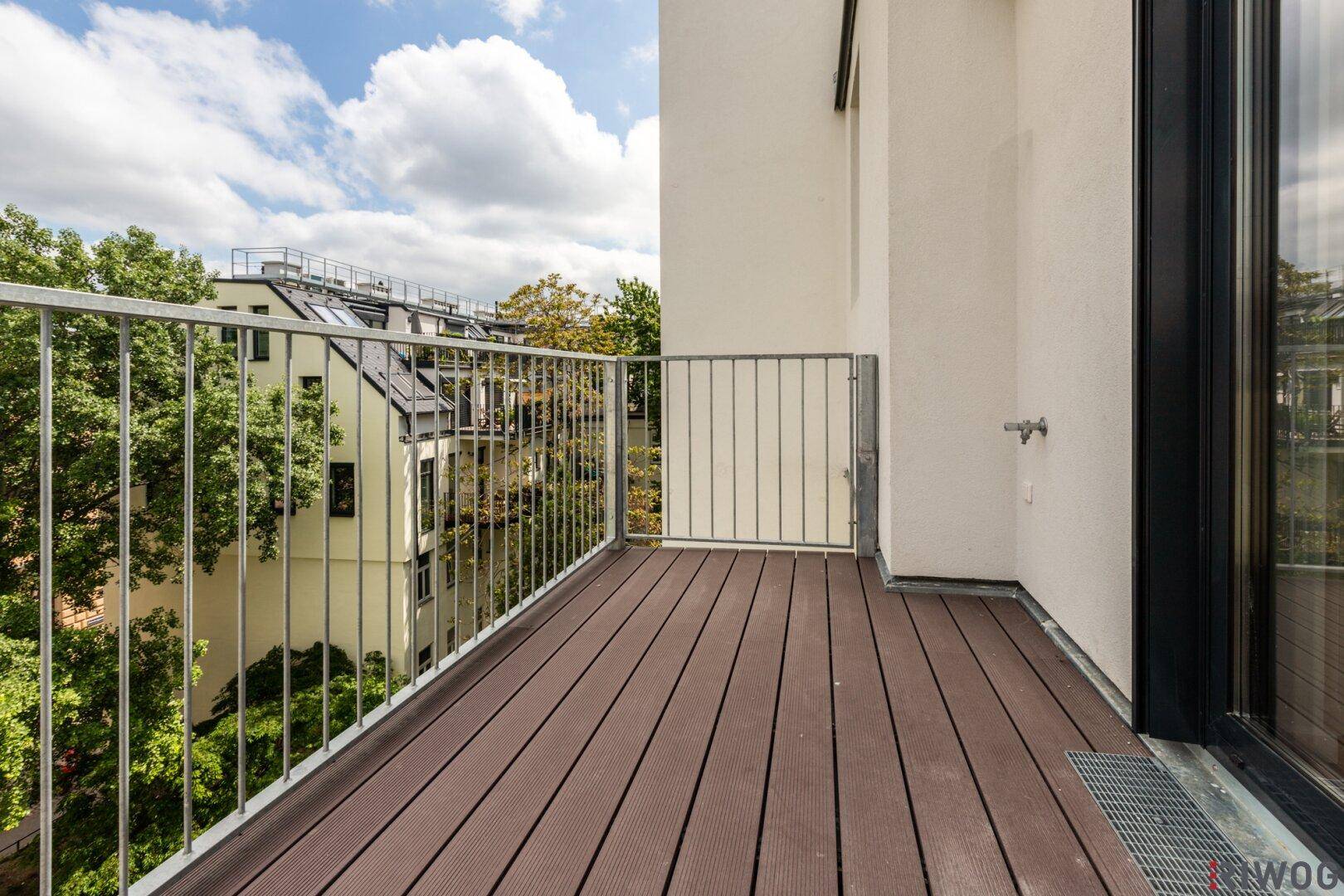 Geräumiger Balkon mit Holzdielen und Blick auf grüne Bäume und Nachbargebäude.