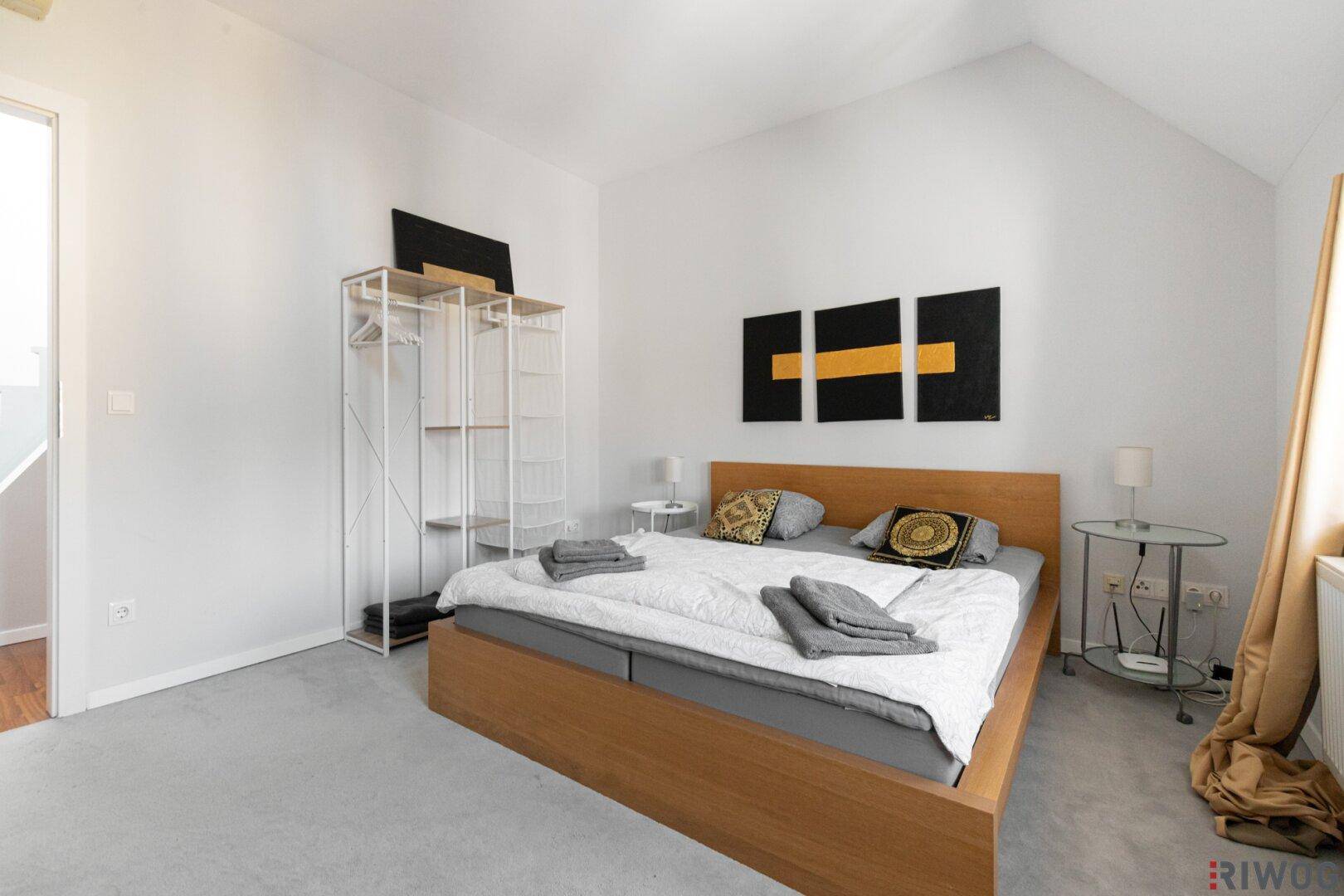 Schlafzimmer mit Holzbett, offenem Kleiderschrank und moderner Wanddekoration.