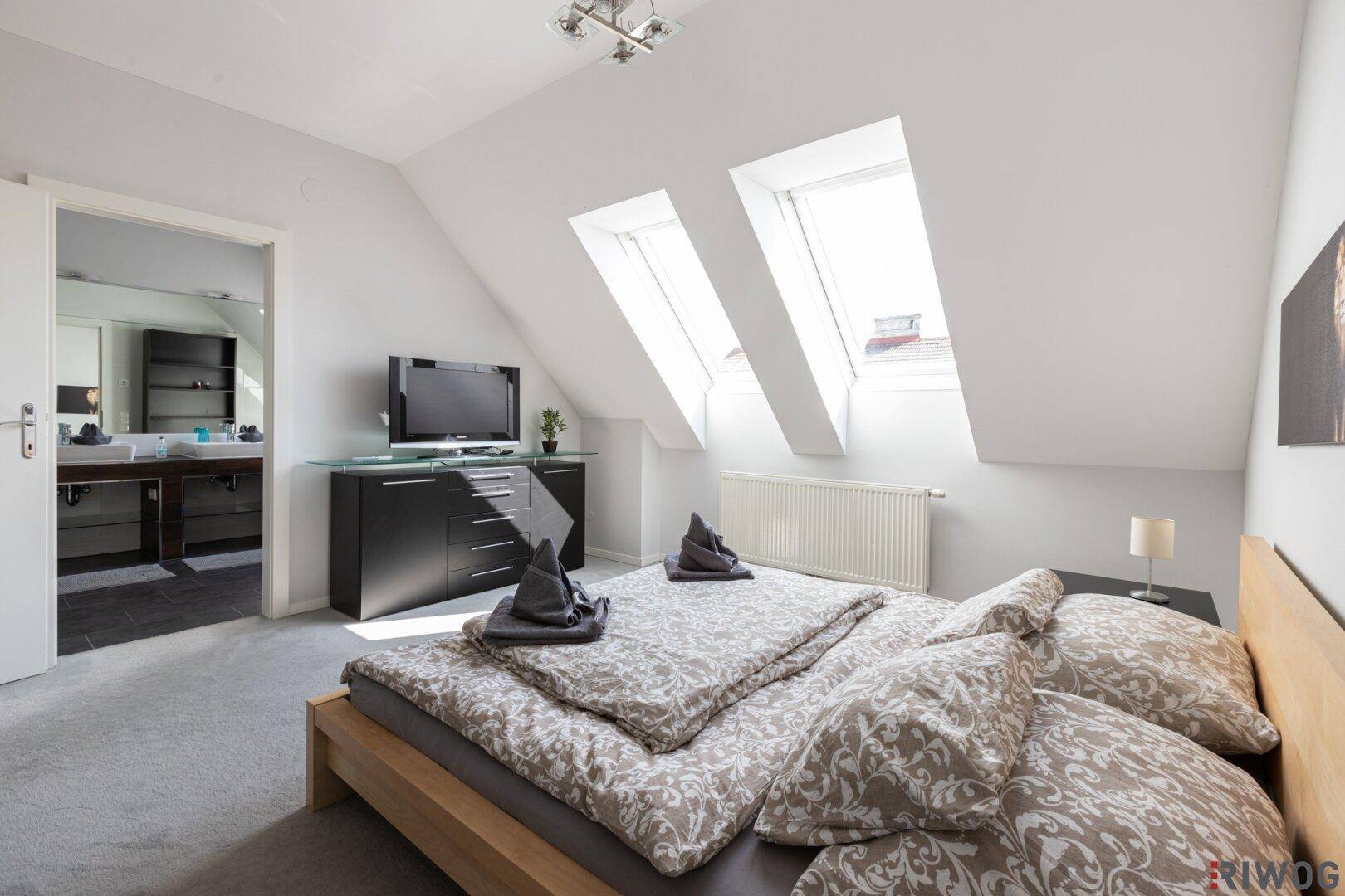 Helles Schlafzimmer im Dachgeschoss mit zwei Dachfenstern und Zugang zum Badezimmer.