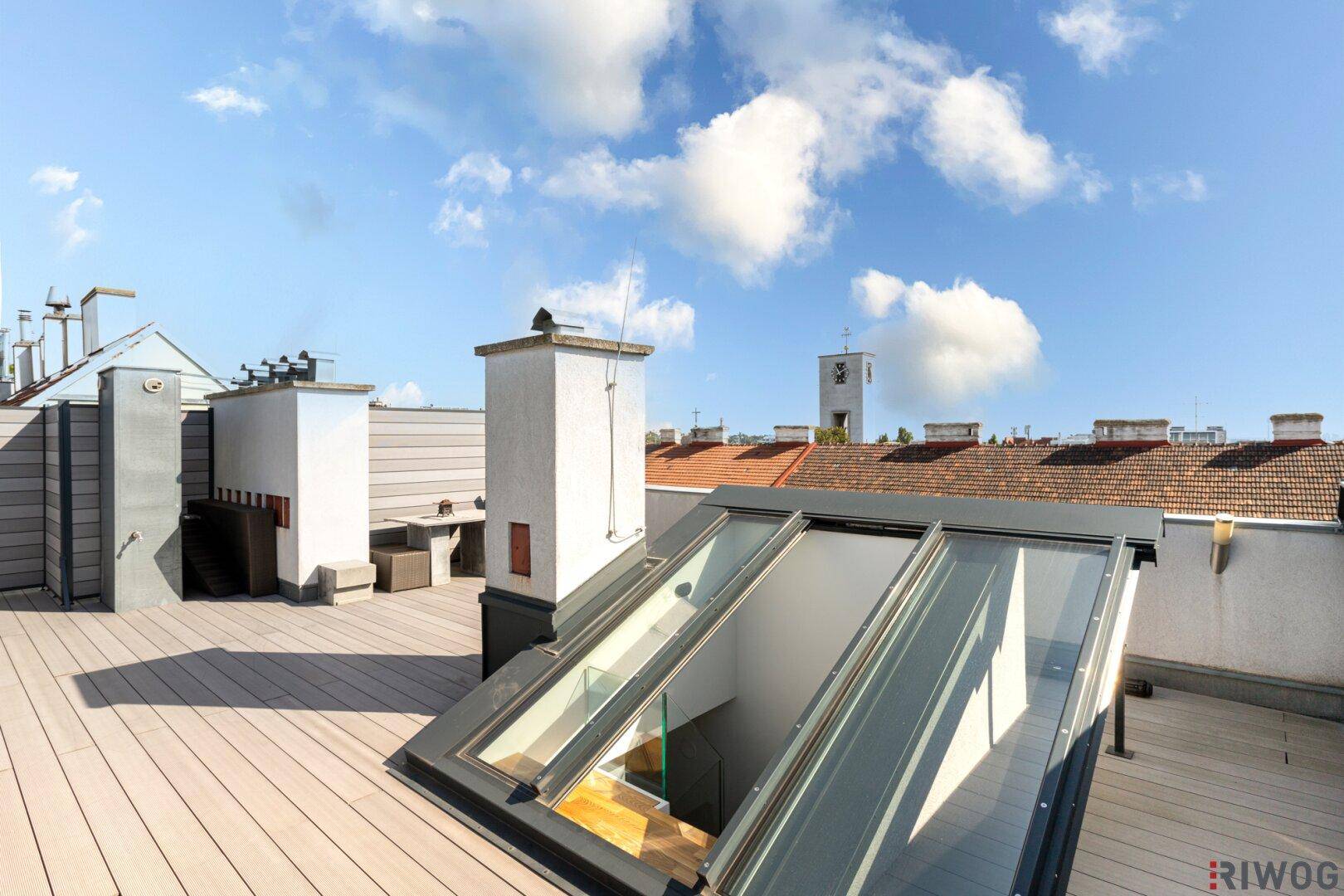 Sonnige Dachterrasse mit Holzdeck, Glasdach-Zugang und Panoramablick über die Dächer der Stadt.