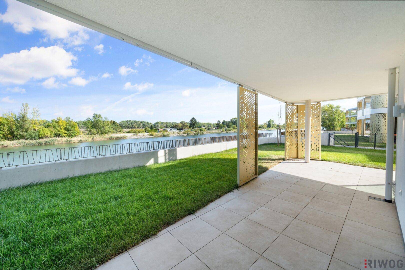 Überdachte Terrasse mit Fliesenboden, Rasenfläche und direktem Blick auf das Wasser.