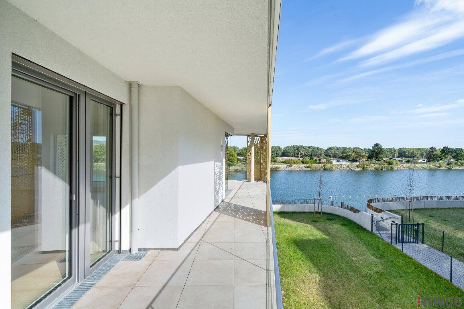 Schmaler Balkon mit Fliesenboden, Glasgeländer und Blick auf den See und eine Grünfläche.