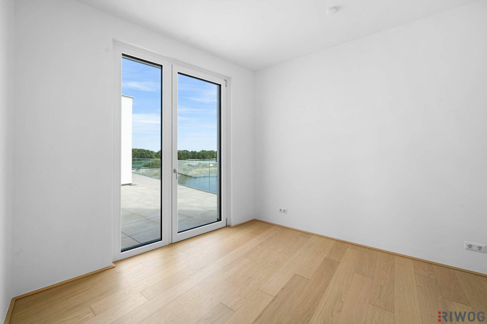 Freundliches Zimmer mit Holzboden und großen Fenstern, die viel Tageslicht hereinlassen.