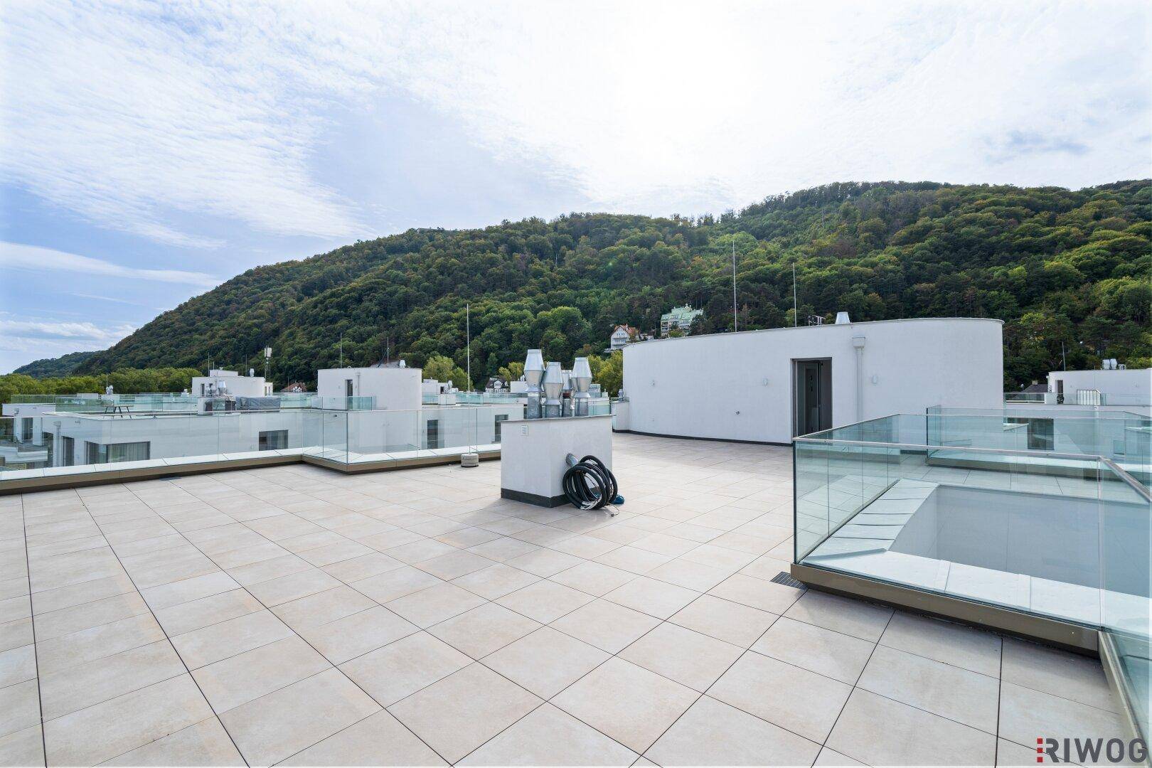 Große Dachterrasse mit Fliesenboden und Glasgeländer, umgeben von grünen Hügeln.