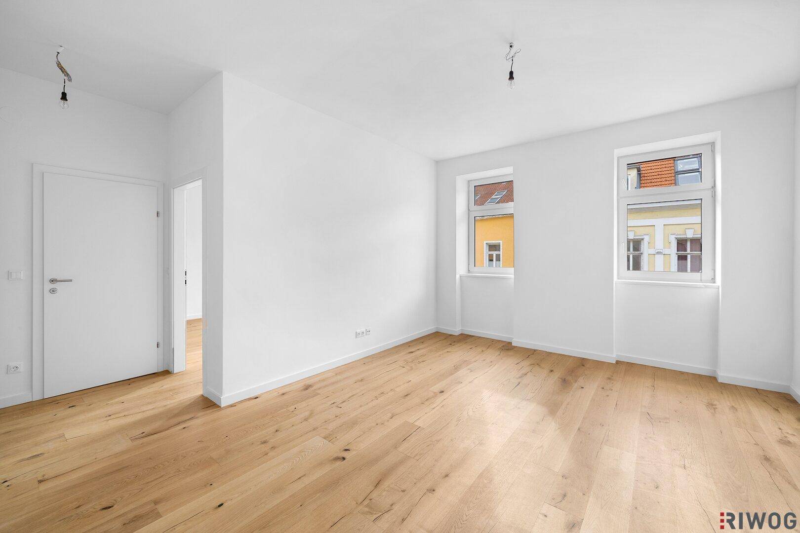 Geräumiger, heller Raum mit Holzfußboden und zwei Fenstern mit Blick auf die umliegenden Gebäude.