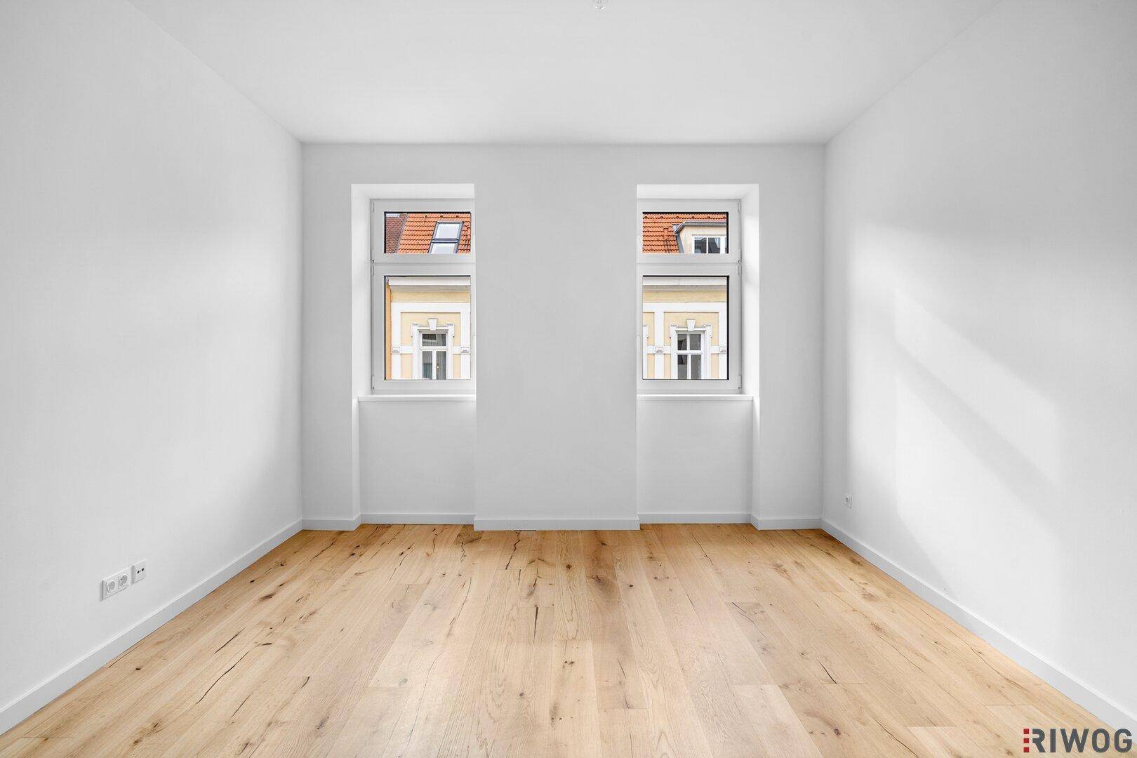 Heller Raum mit Holzfußboden und zwei Fenstern, die viel Tageslicht in den Wohnbereich bringen.