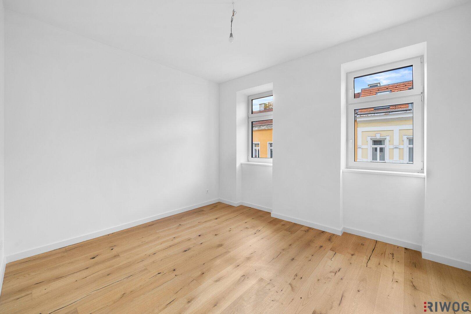 Lichtdurchfluteter Raum mit Holzfußboden und zwei Fenstern, ideal für ein Schlafzimmer oder Büro.
