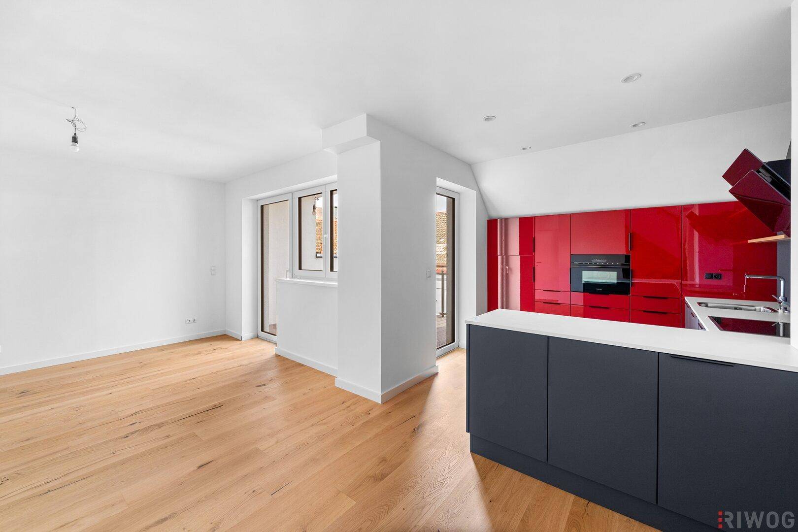 Heller Wohnbereich mit Holzboden und moderner, offener Küche in Rot und Grau, mit Zugang zur Loggia.