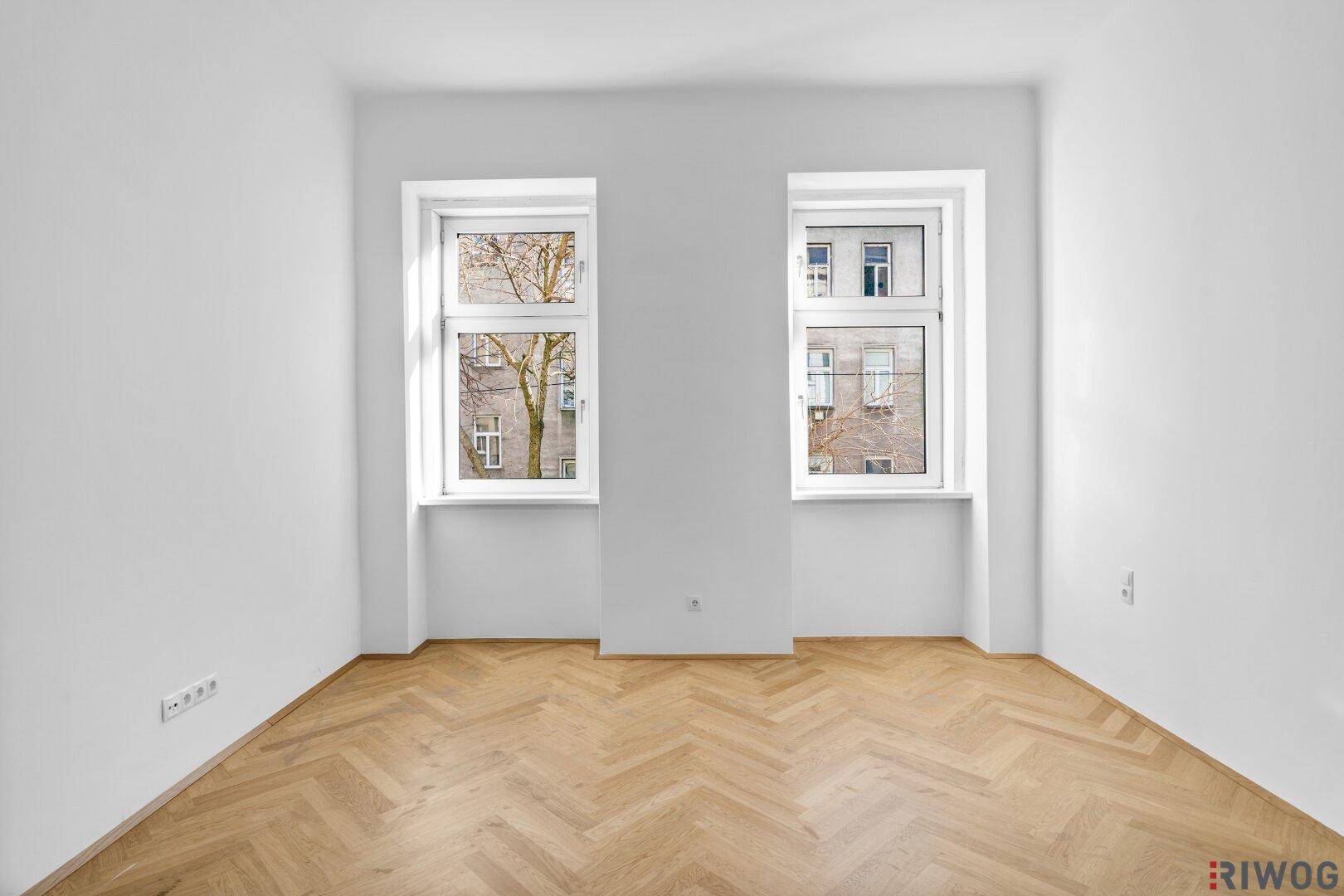 Heller Raum mit zwei Fenstern und Fischgrätparkett, bietet viel Tageslicht und Ausblick.