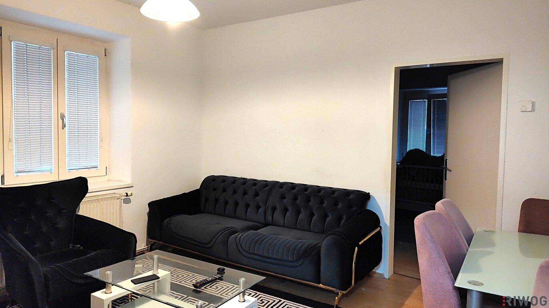 Gemütliches Wohnzimmer mit dunklem Samtsofa, Couchtisch und hellem Fenster mit Jalousien.