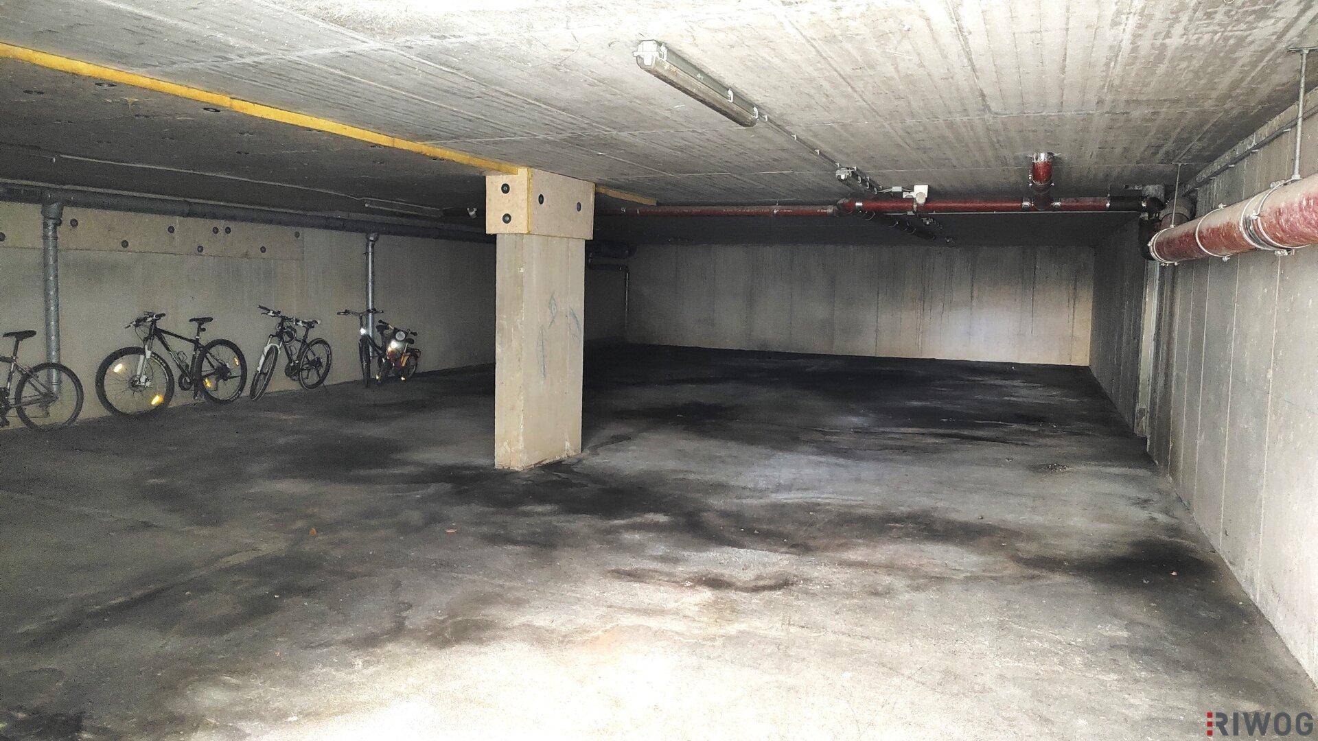 Tiefgarage mit Betonwänden und -boden, Fahrrädern und sichtbaren Leitungen an der Decke.