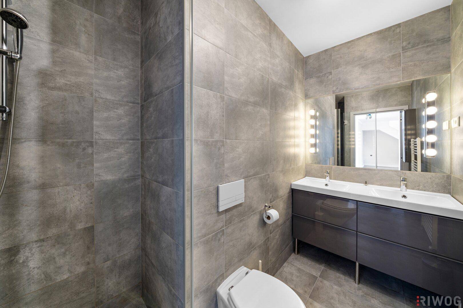 Modernes Badezimmer mit grauen Fliesen, Doppelwaschbecken und großer Dusche.