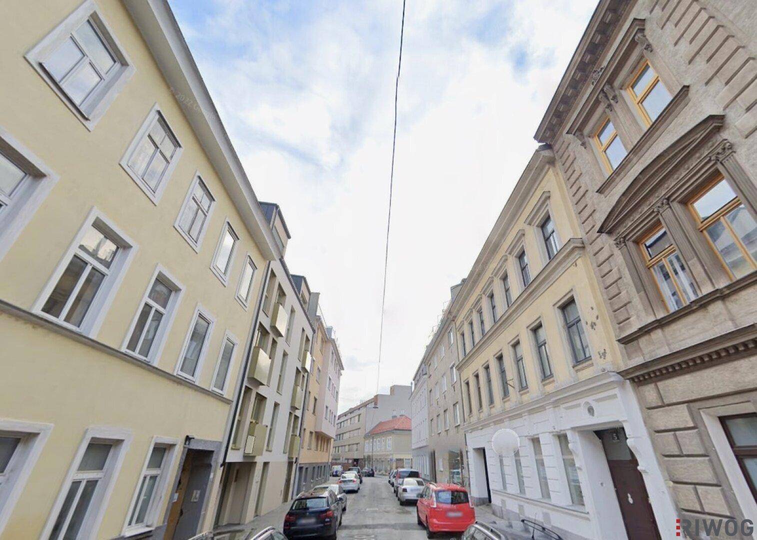 Blick entlang einer städtischen Straße mit mehrstöckigen Gebäuden und parkenden Autos unter bewölktem Himmel.
