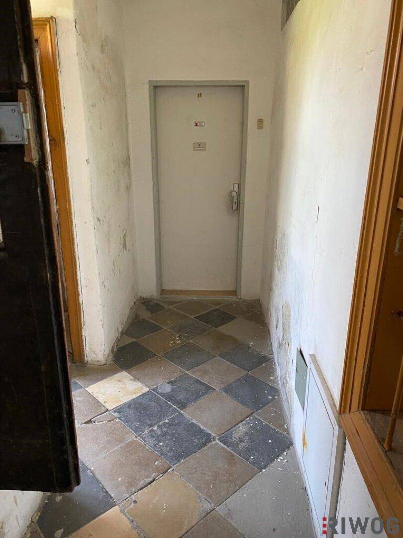Eingangsbereich mit abgenutzten, karierten Fliesen und schlichten Wänden, die Renovierungsbedarf zeigen.
