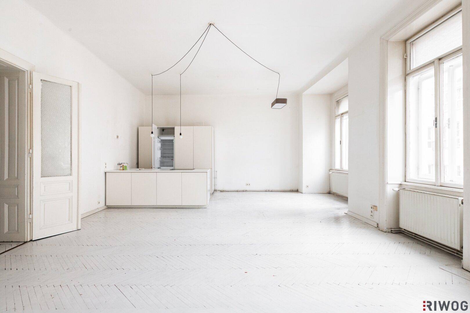 Geräumiger, heller Wohnbereich mit integrierter Küchenzeile und hohen Fenstern, typisch für einen Altbau.