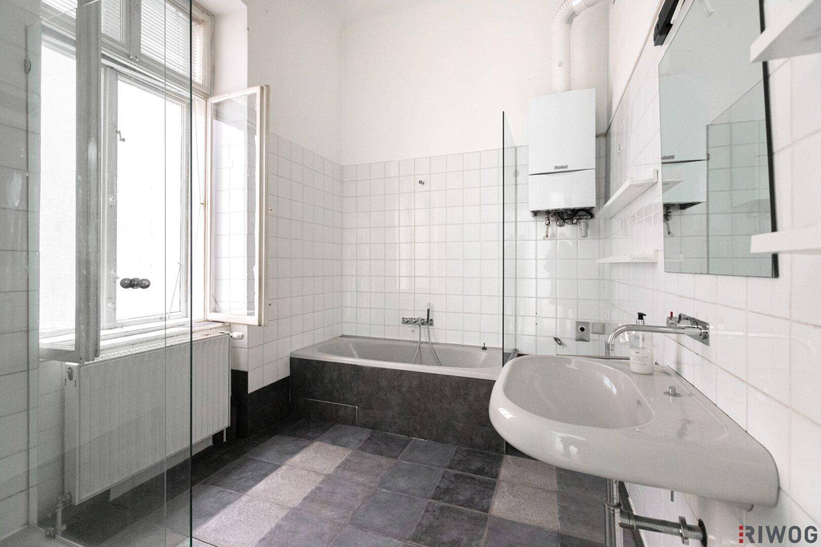 Badezimmer mit Badewanne, Dusche und Fenster, ausgestattet mit weißen Fliesen und dunklem Boden.