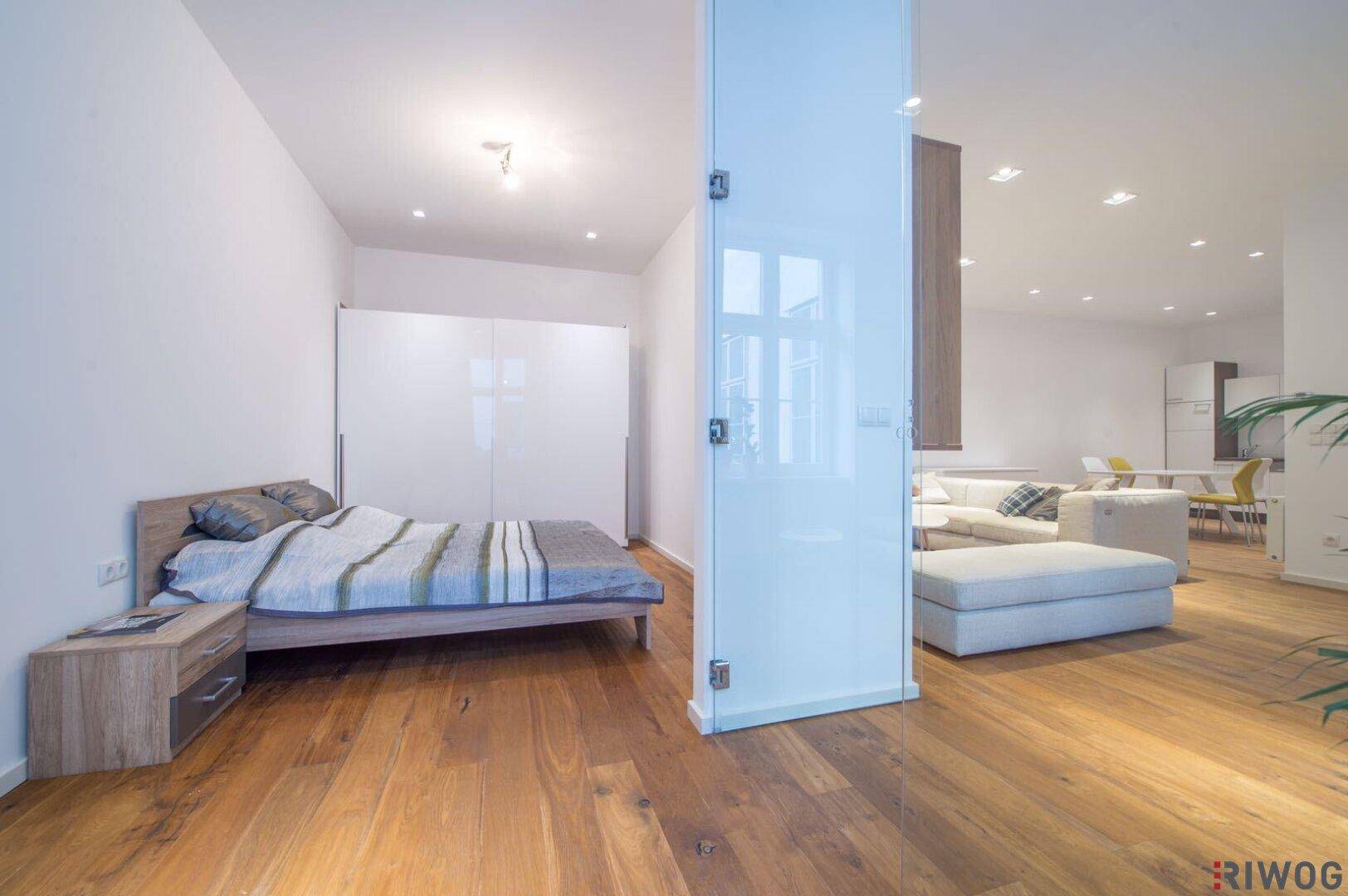 Helles Schlafzimmer mit Holzboden, großem Bett und weißen Einbauschränken, getrennt durch eine Glaswand.