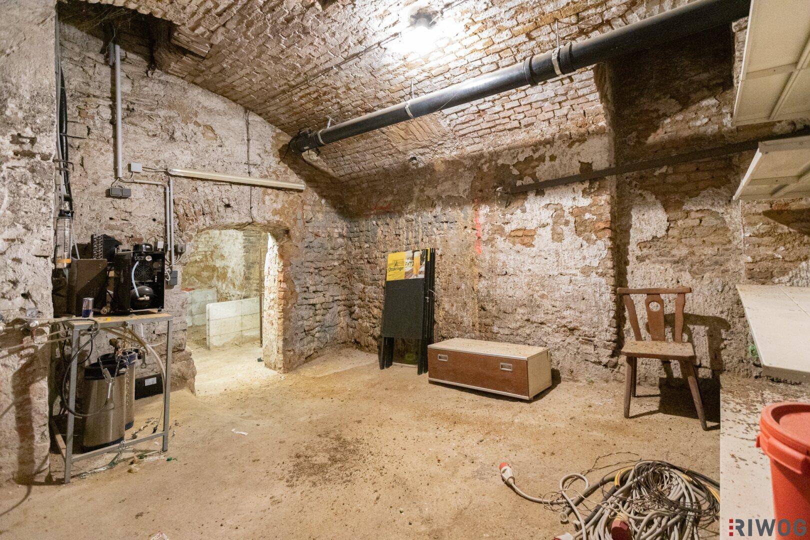 Rustikaler Kellerraum mit unverputzten Ziegelwänden, Gewölbedecke und Rohren an der Decke.