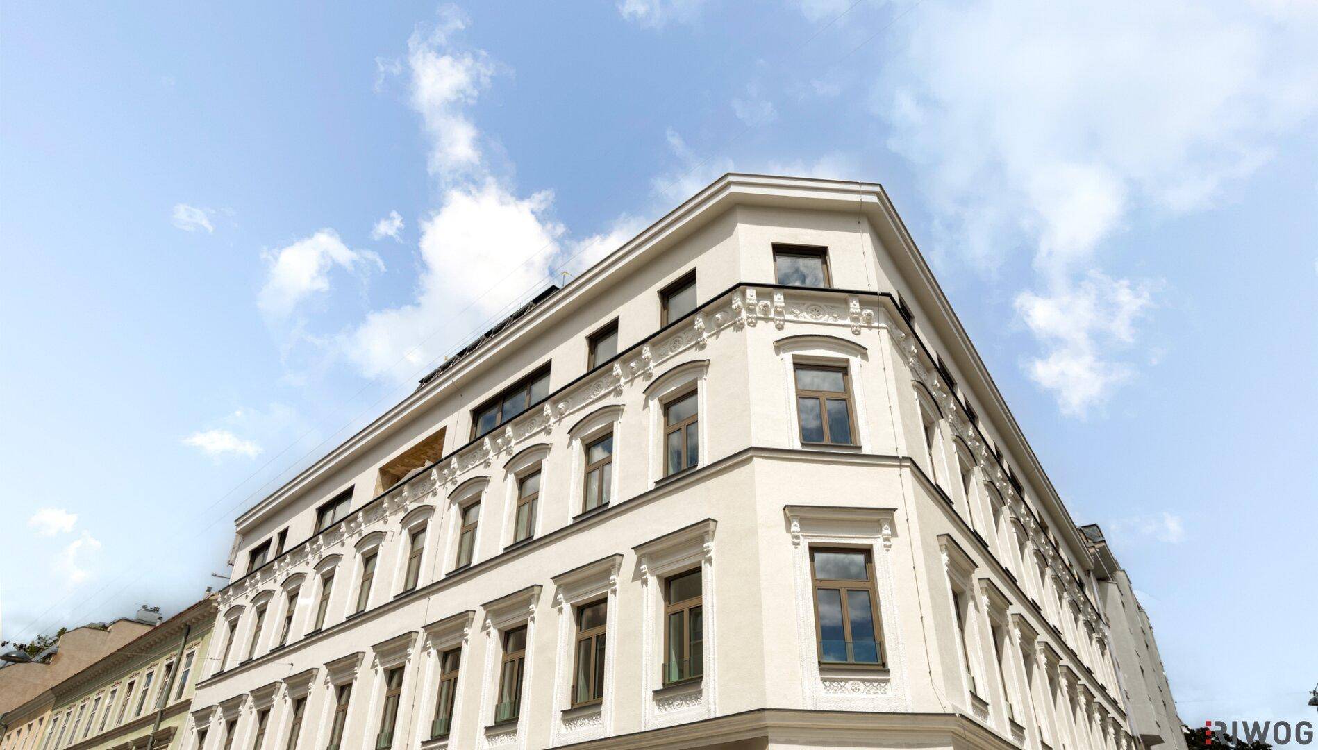 Helle Fassade eines Eckgebäudes mit klassischen Verzierungen und modernen Fenstern unter blauem Himmel.