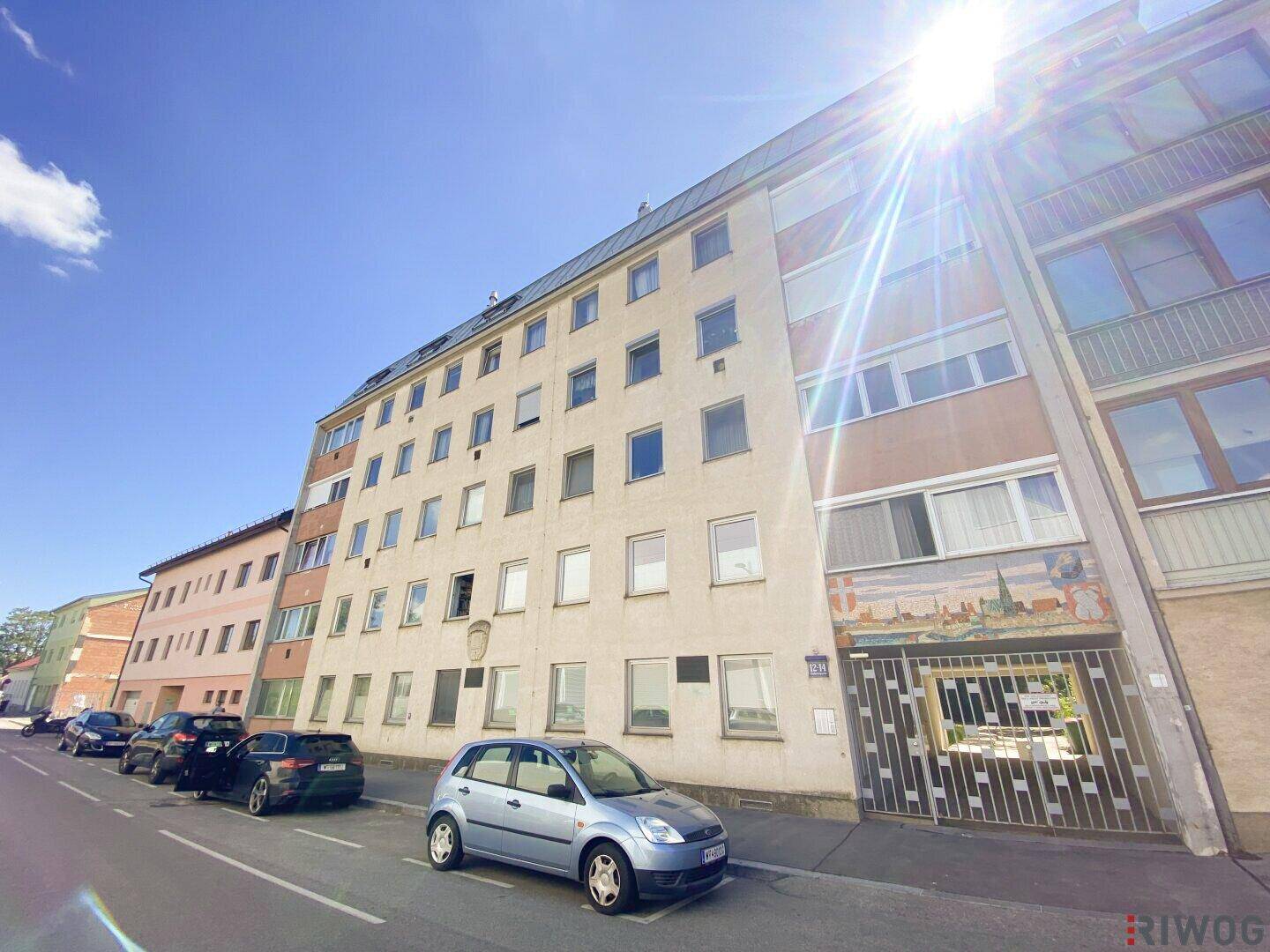 Mehrfamilienhaus mit mehreren Etagen und geparkten Autos davor, unter strahlend blauem Himmel.