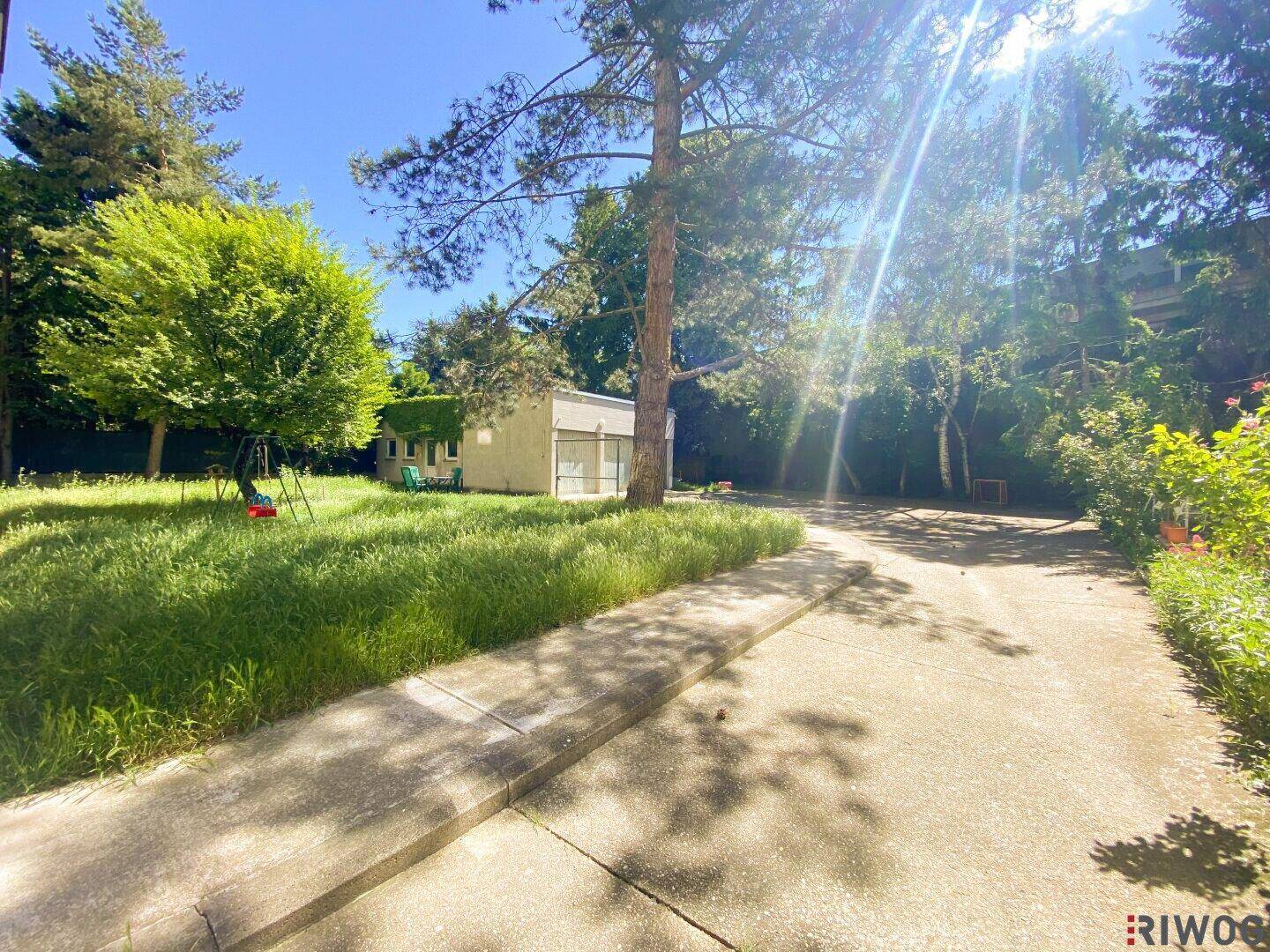 Weitläufige Gartenanlage mit altem Baumbestand, Rasenflächen und einem kleinen Gebäude im Hintergrund.