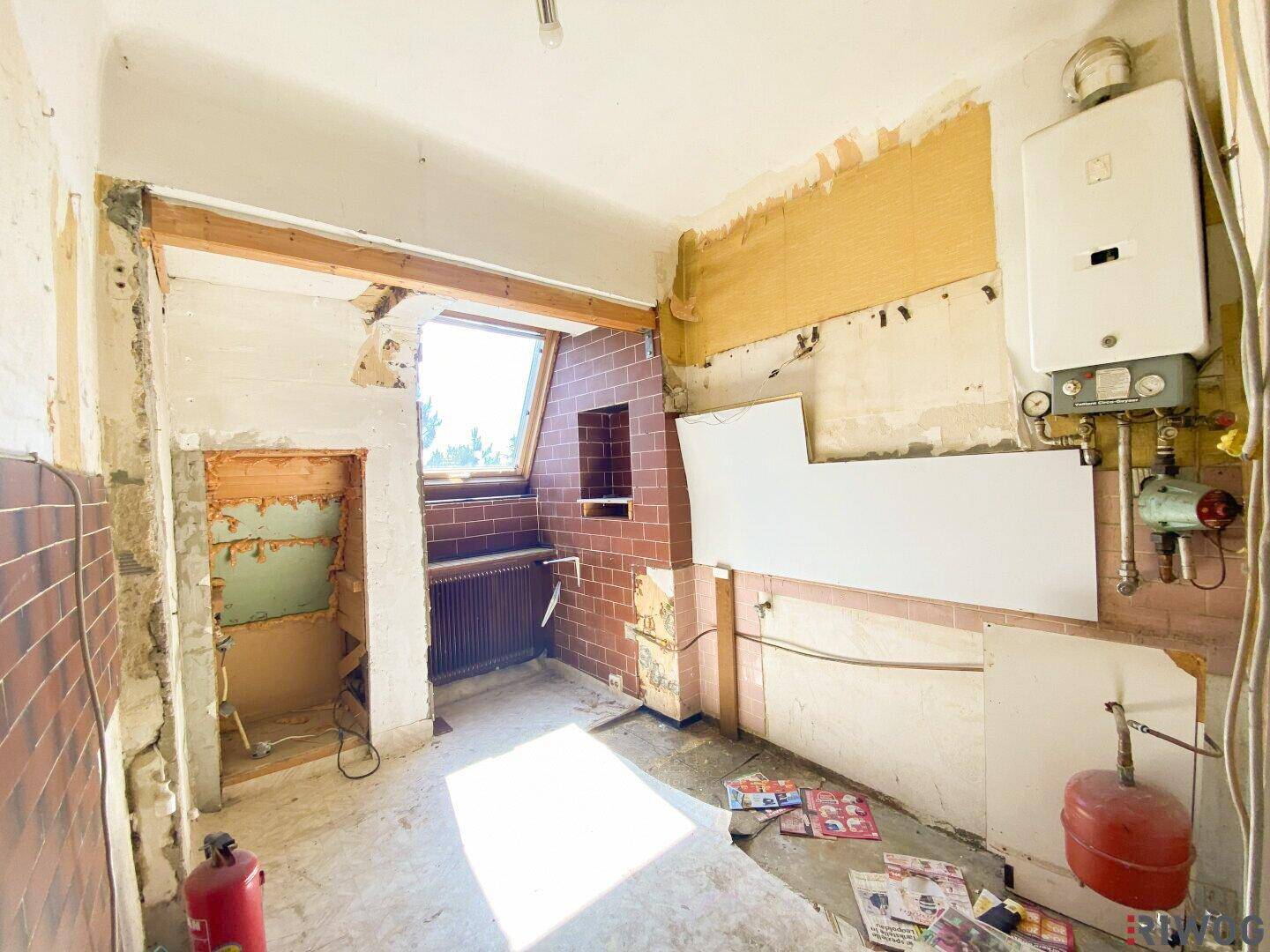 Raum im Abrisszustand mit freigelegten Wänden, Heizungsanlage und einem Dachfenster, das Licht spendet.