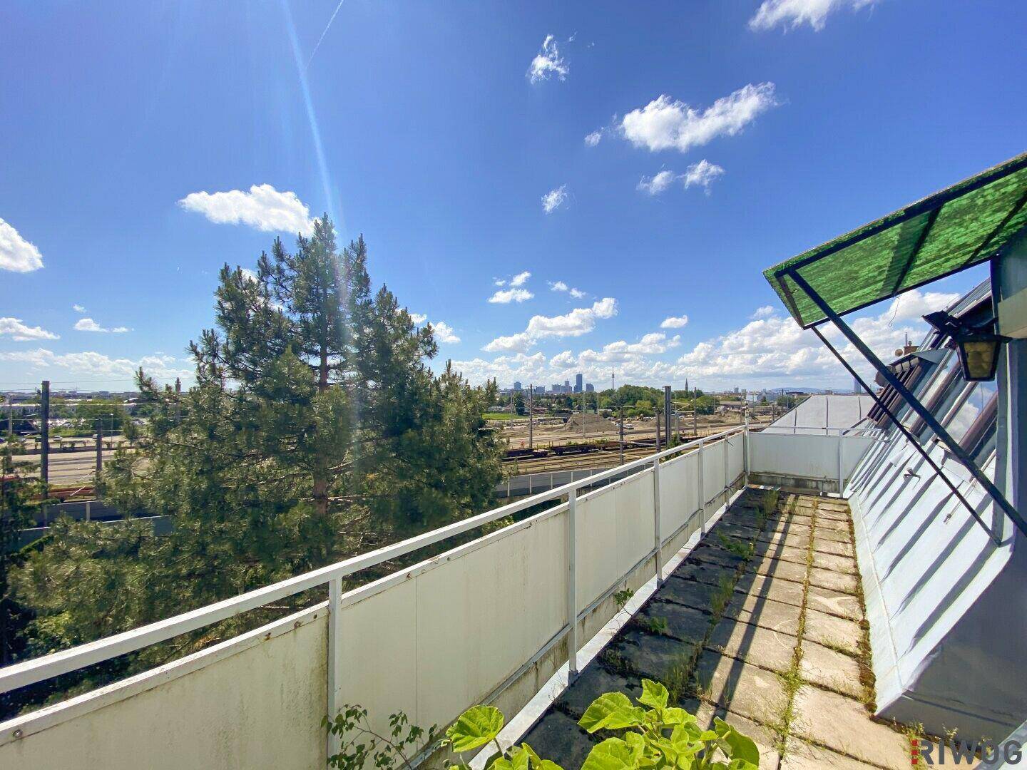 Langer Balkon mit Blick auf Bahngleise und die Skyline der Stadt unter blauem Himmel.