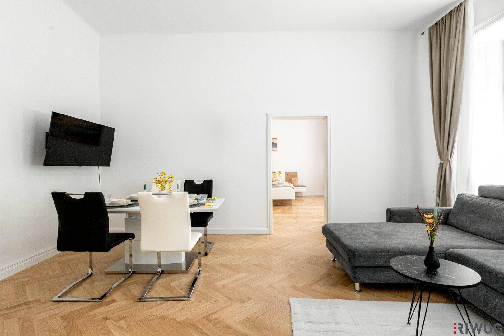Heller Wohnbereich mit Esstisch, Sofa und Zugang zu einem weiteren Zimmer.