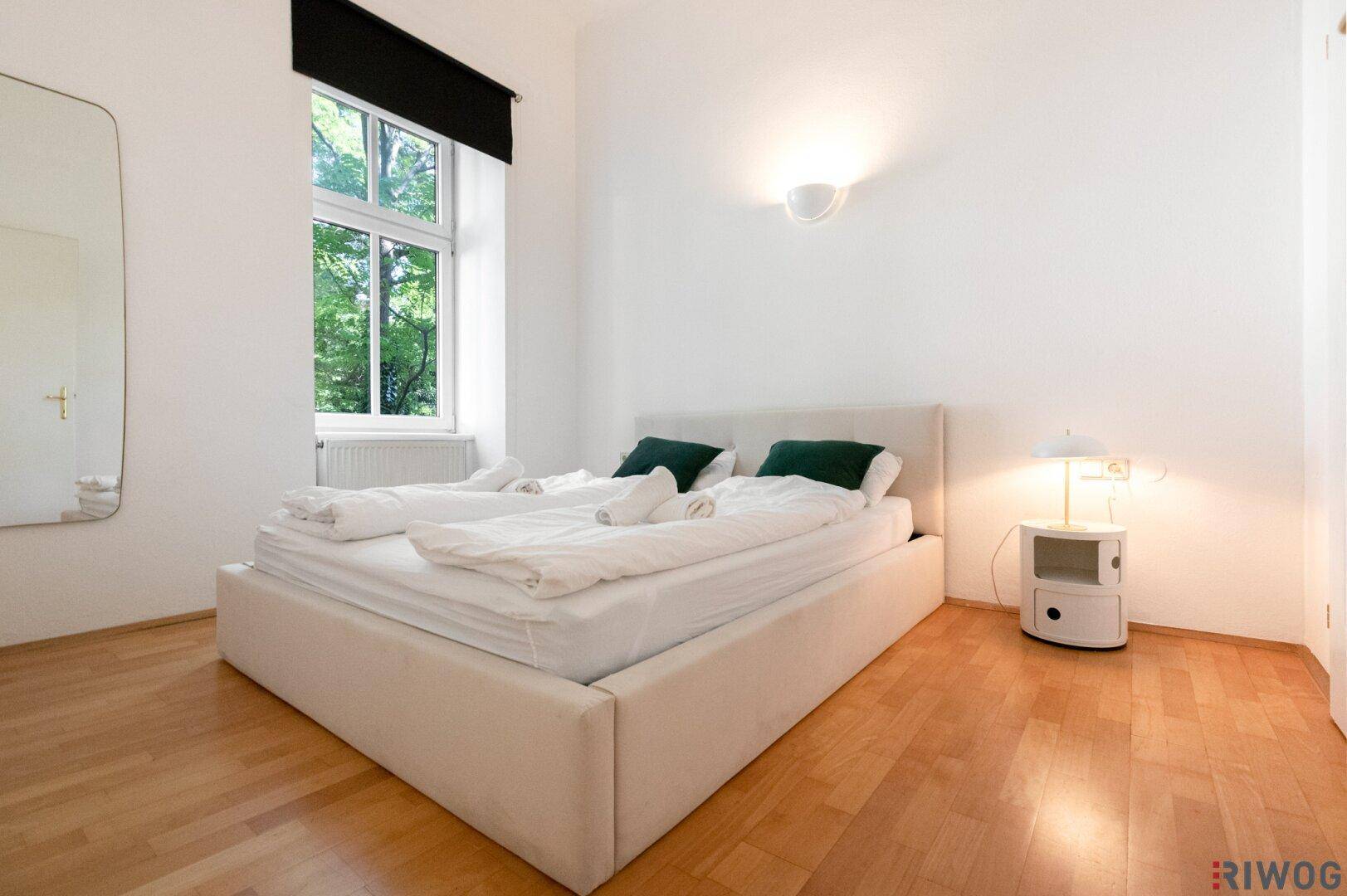 Gemütliches Schlafzimmer mit weißem Bett, Parkettboden und viel Tageslicht.