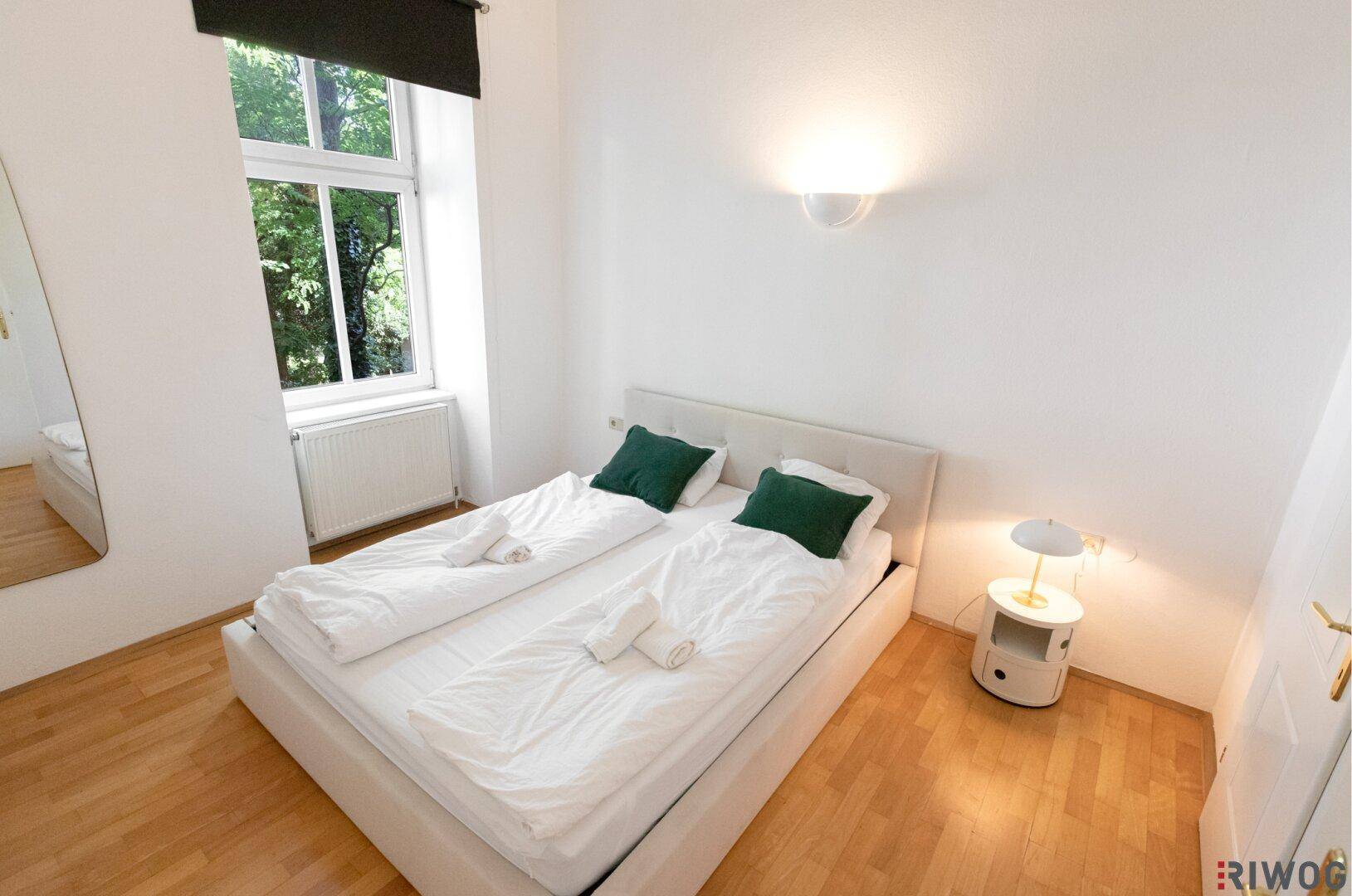 Helles Schlafzimmer mit Doppelbett, Nachttisch und Fenster mit Blick ins Grüne.