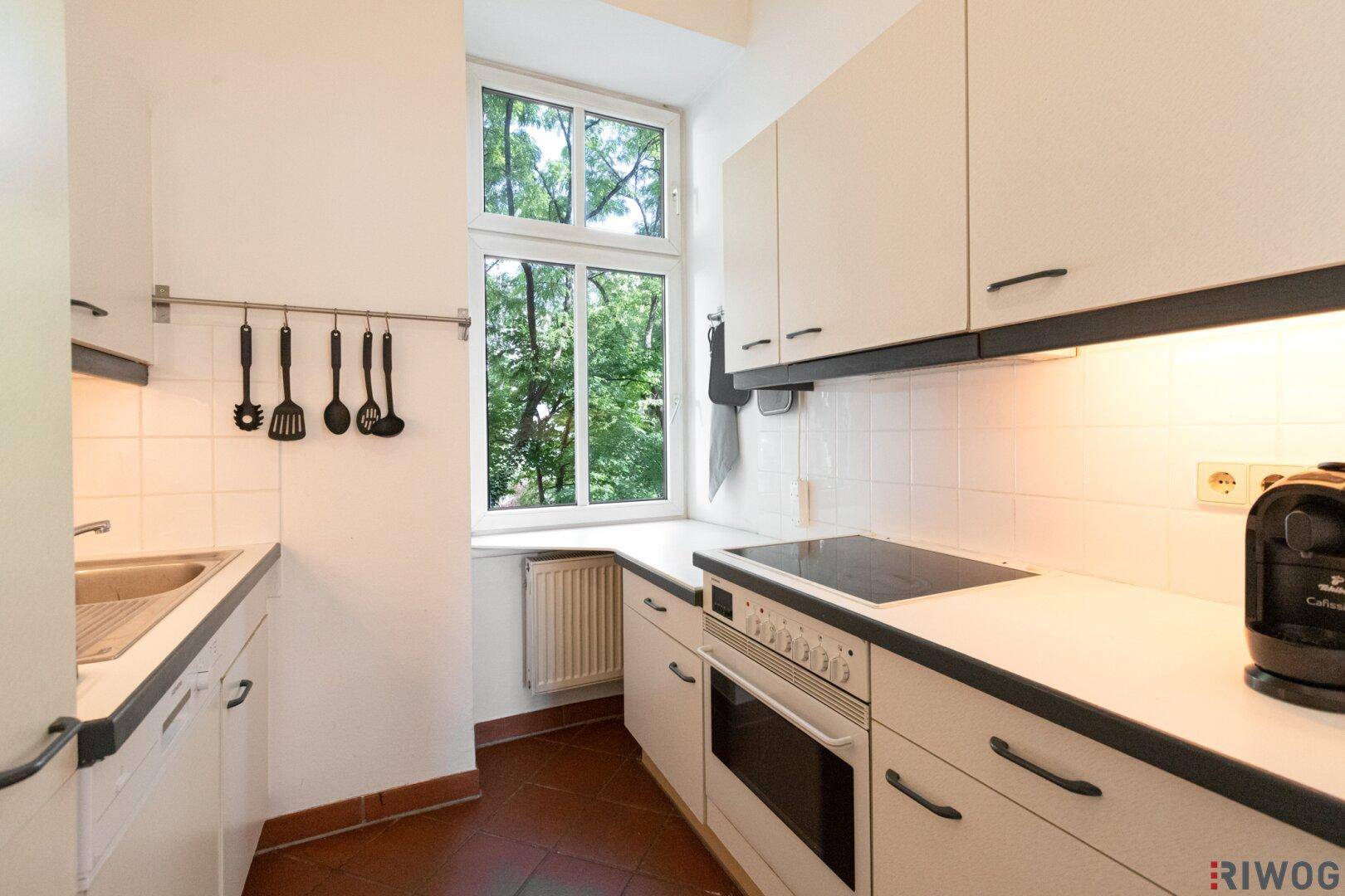 Kleine Küche mit weißen Einbauschränken, Elektroherd und Fenster mit Blick ins Grüne.
