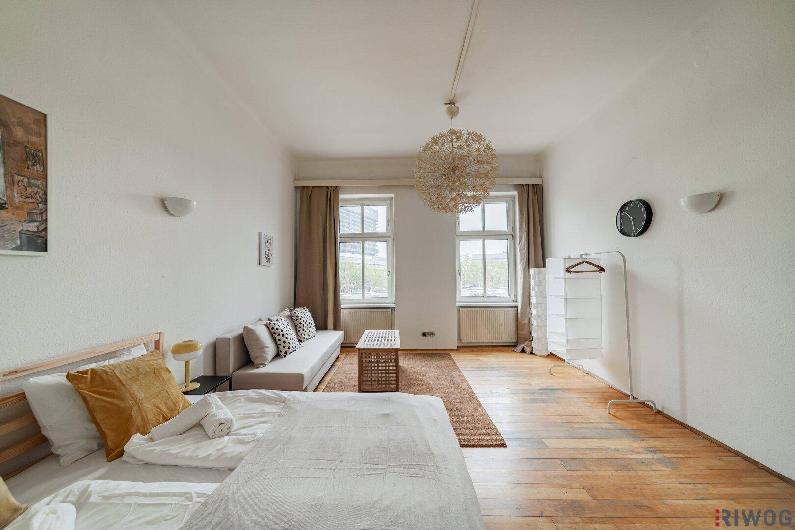 Helles Zimmer mit Holzboden, Bett, Sofa und großem Fenster, ideal als Wohn- oder Schlafbereich.
