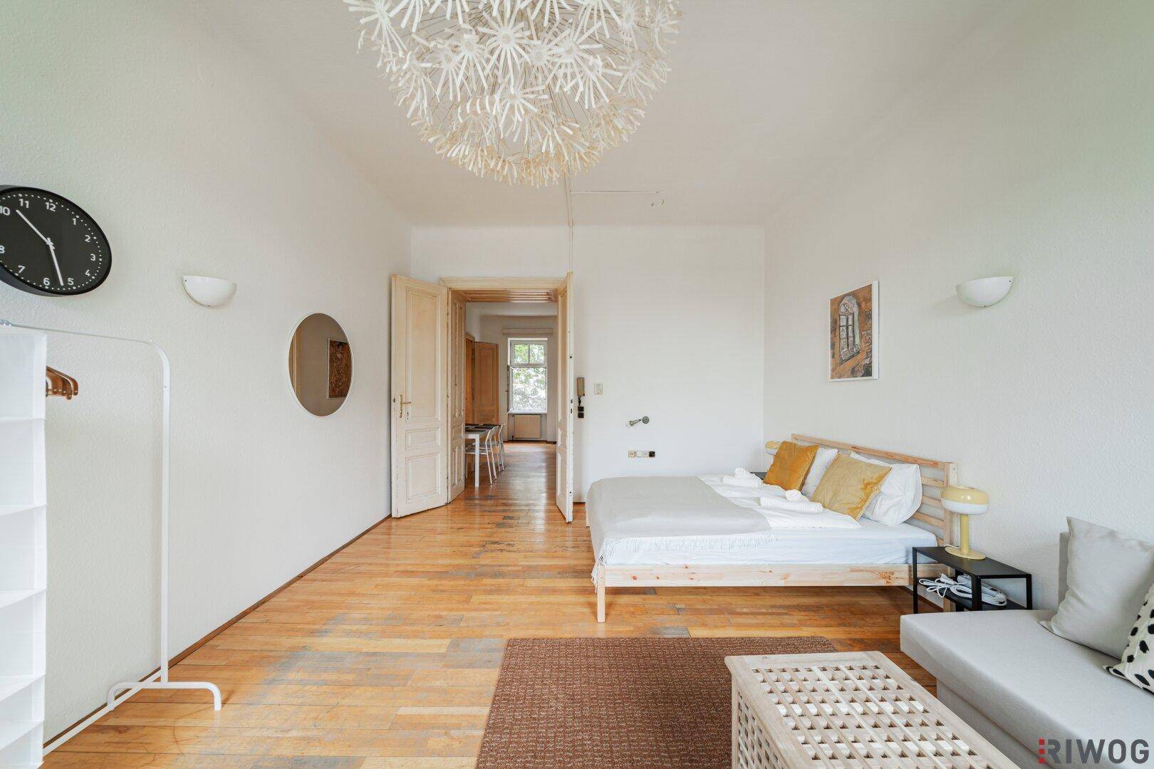 Wohnzimmer mit Holzboden und Zugang zu einem weiteren Raum, ausgestattet mit Bett und Sofa.