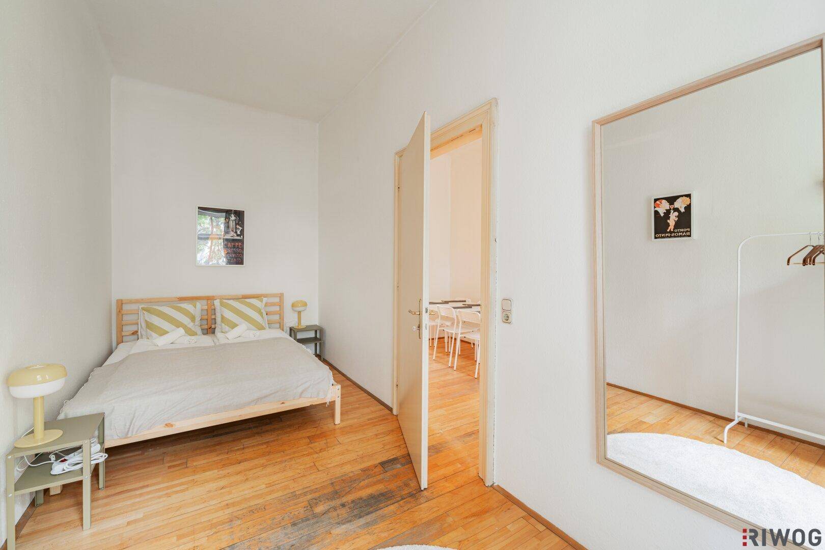 Schlafzimmer mit Holzboden, großem Spiegel und offener Tür zu einem angrenzenden Raum.