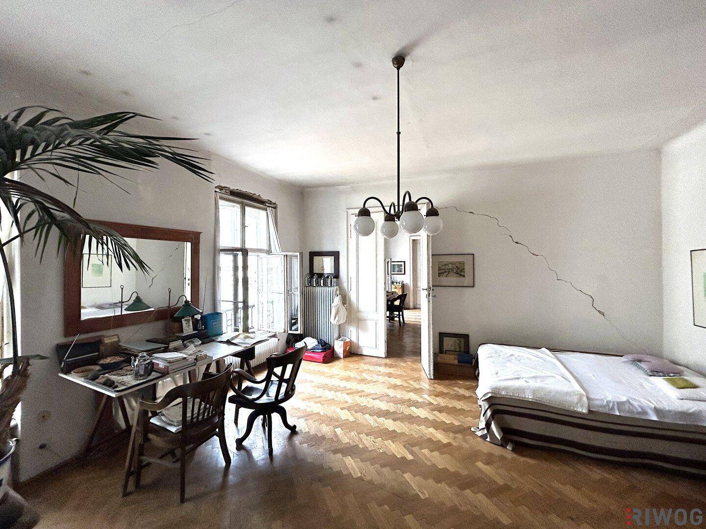 Helles Wohnzimmer mit Parkettboden, Rissen an den Wänden und Blick in einen weiteren Raum.