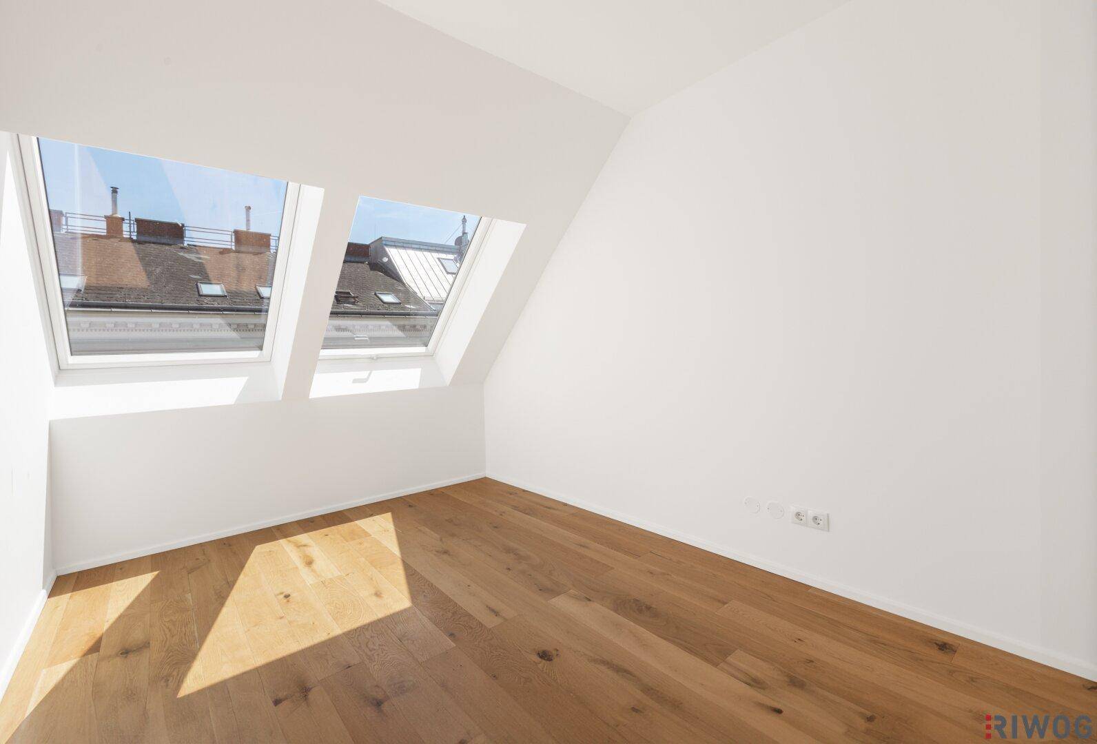 Lichtdurchflutetes Dachzimmer mit Holzboden und Blick auf die umliegenden Dächer durch die Dachfenster.