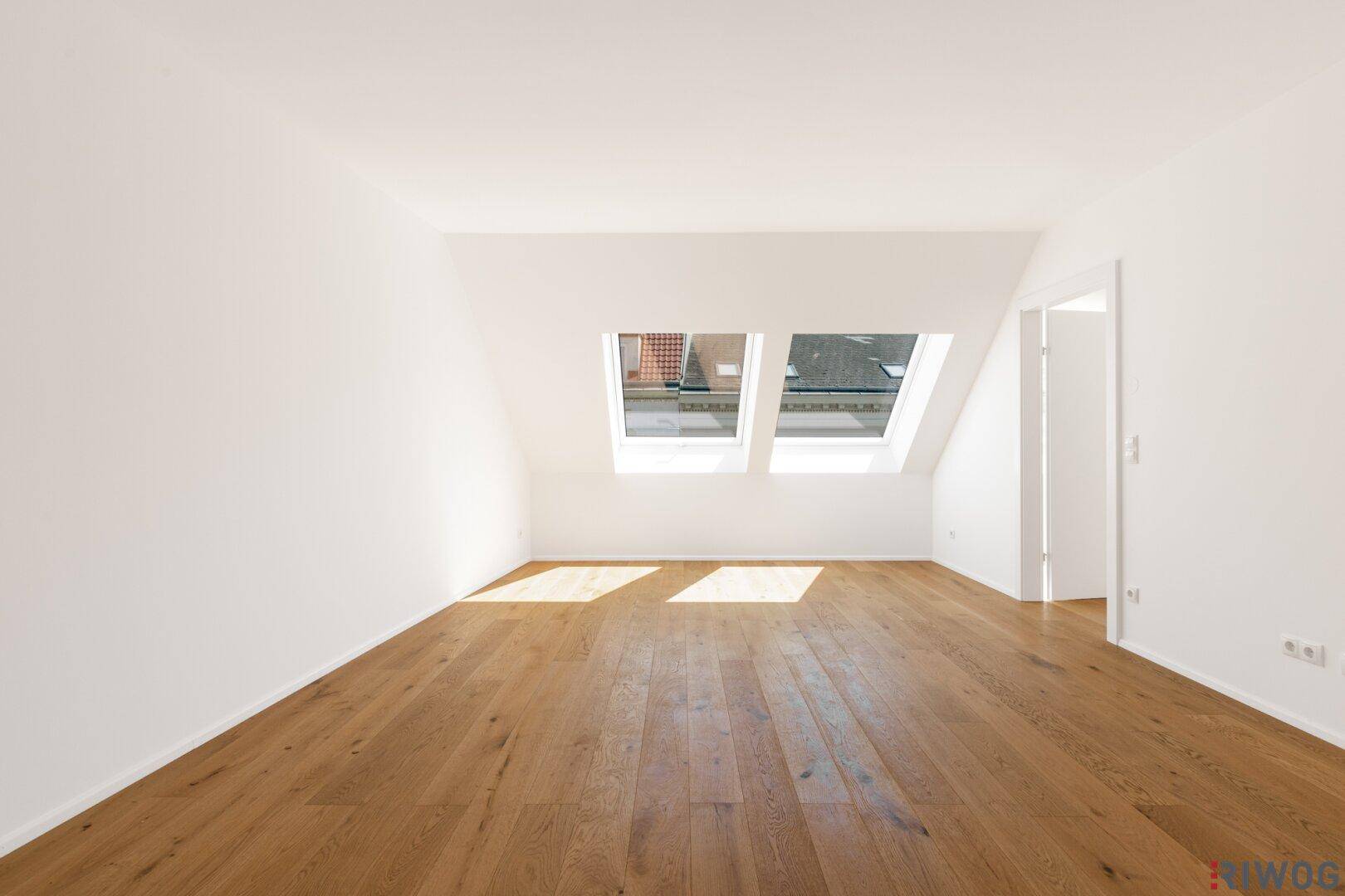 Heller Raum im Dachgeschoss mit Holzboden und zwei Dachfenstern, durch die Sonnenlicht fällt.