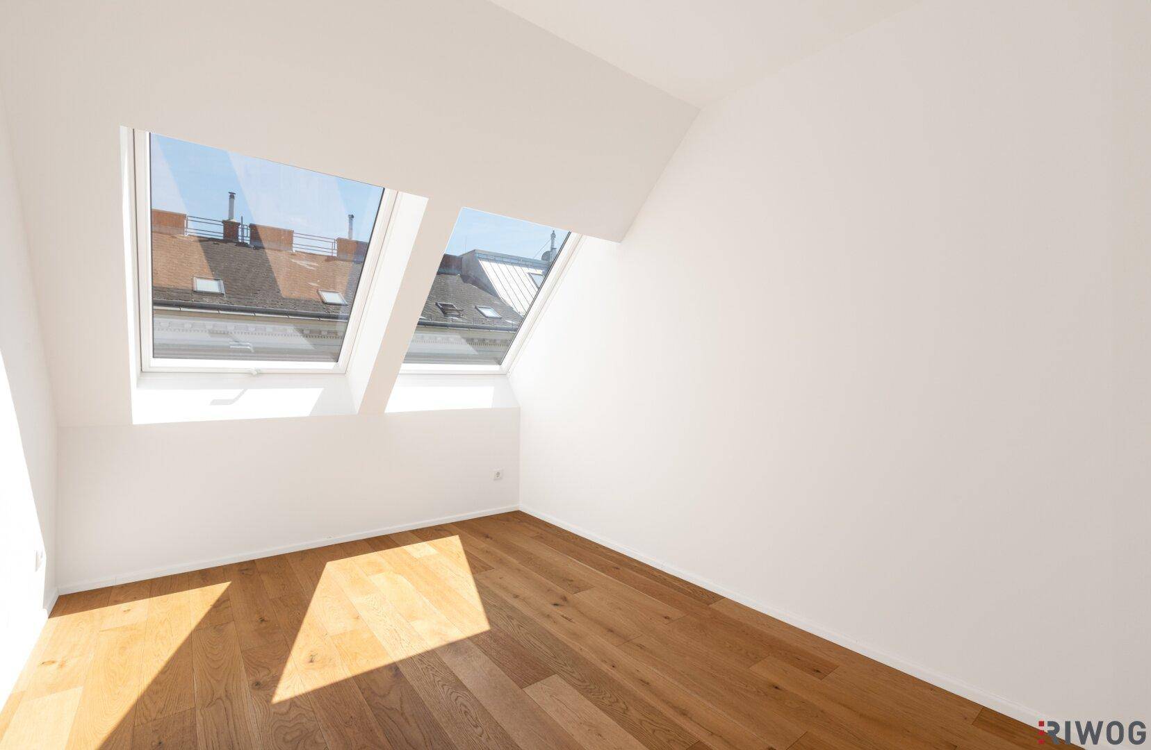 Geräumiger Raum im Dachgeschoss mit schrägen Wänden, Holzboden und zwei großen Dachfenstern für viel Helligkeit.
